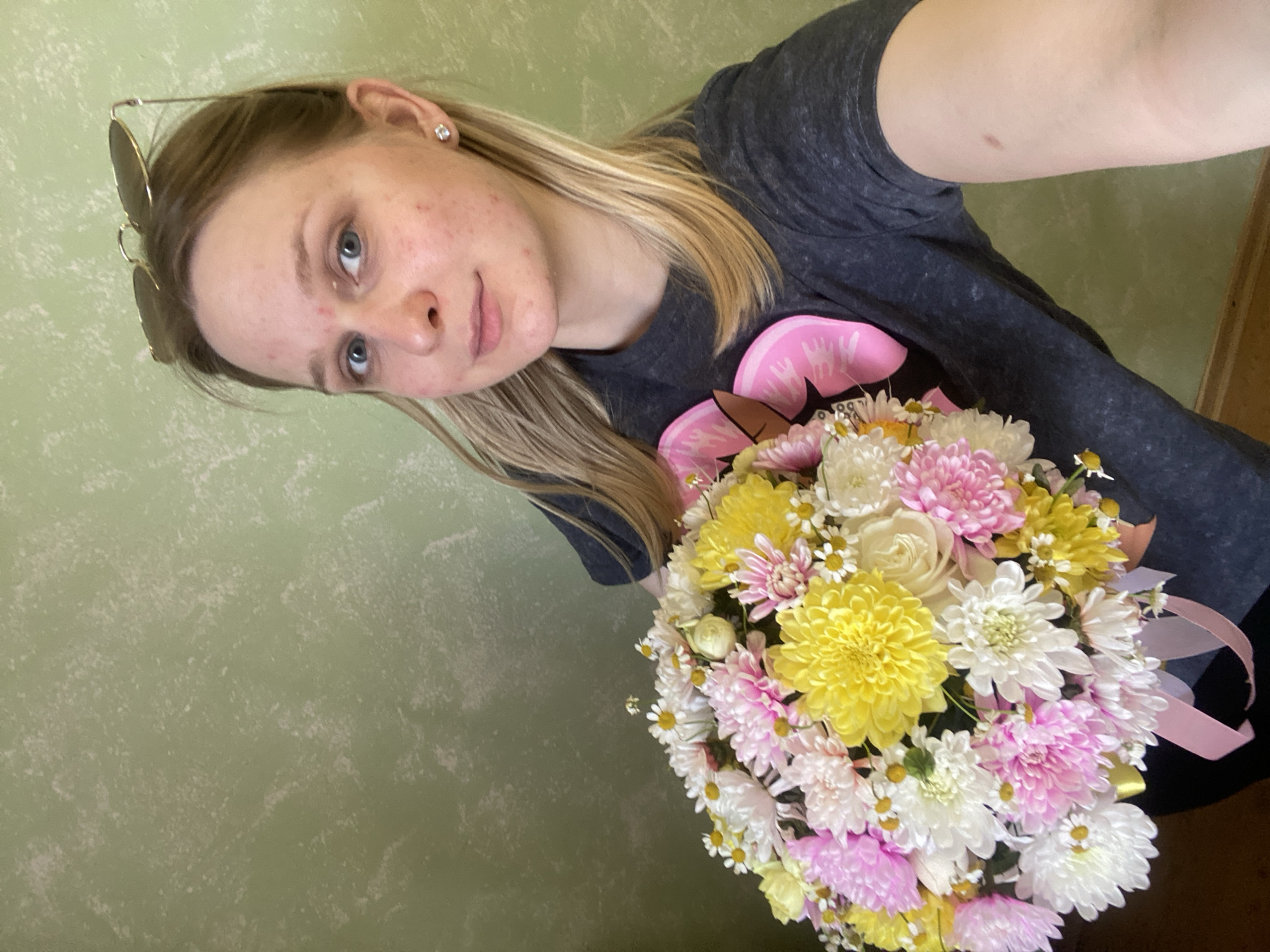 The image depicts a close-up, upward-angled selfie of a young Caucasian female, approximately in her late teens or early twenties, holding a large floral bouquet indoors in the city of Unknown, Unknown. She has long, straight, light blonde hair and visible acne blemishes on her forehead, nose, cheeks, and chin. Her eyes are blue, and she exhibits a slight, closed-mouth smile. Gold-rimmed aviator-style sunglasses are perched on top of her head. A clear, round stud earring is visible in her right ear. She is wearing a dark grey, short-sleeved t-shirt with a portion of a faded pink graphic design visible on the chest.
The focal point of the image is the large, spherical bouquet she is holding, positioned centrally and extending across the lower half of the frame. The bouquet is composed of various flowers, predominantly multi-petaled chrysanthemums or similar flowers in shades of vibrant yellow, light pink, and white. Numerous small, white daisy-like flowers with yellow centers are interspersed throughout the arrangement, along with a few visible light green or cream-colored flower buds. A light pink satin ribbon is tied around the base of the bouquet, with loops and trailing ends visible on the lower right.
The background consists of a solid wall painted in a muted, light green color. The wall surface exhibits a distinct, irregular textured finish, characterized by lighter, whitish, mottled patches against the green base. A narrow strip of a dark brown, possibly wooden, surface (like a baseboard or furniture edge) is visible along the bottom right edge of the frame. The lighting is even and soft, suggesting an indoor environment without harsh shadows.