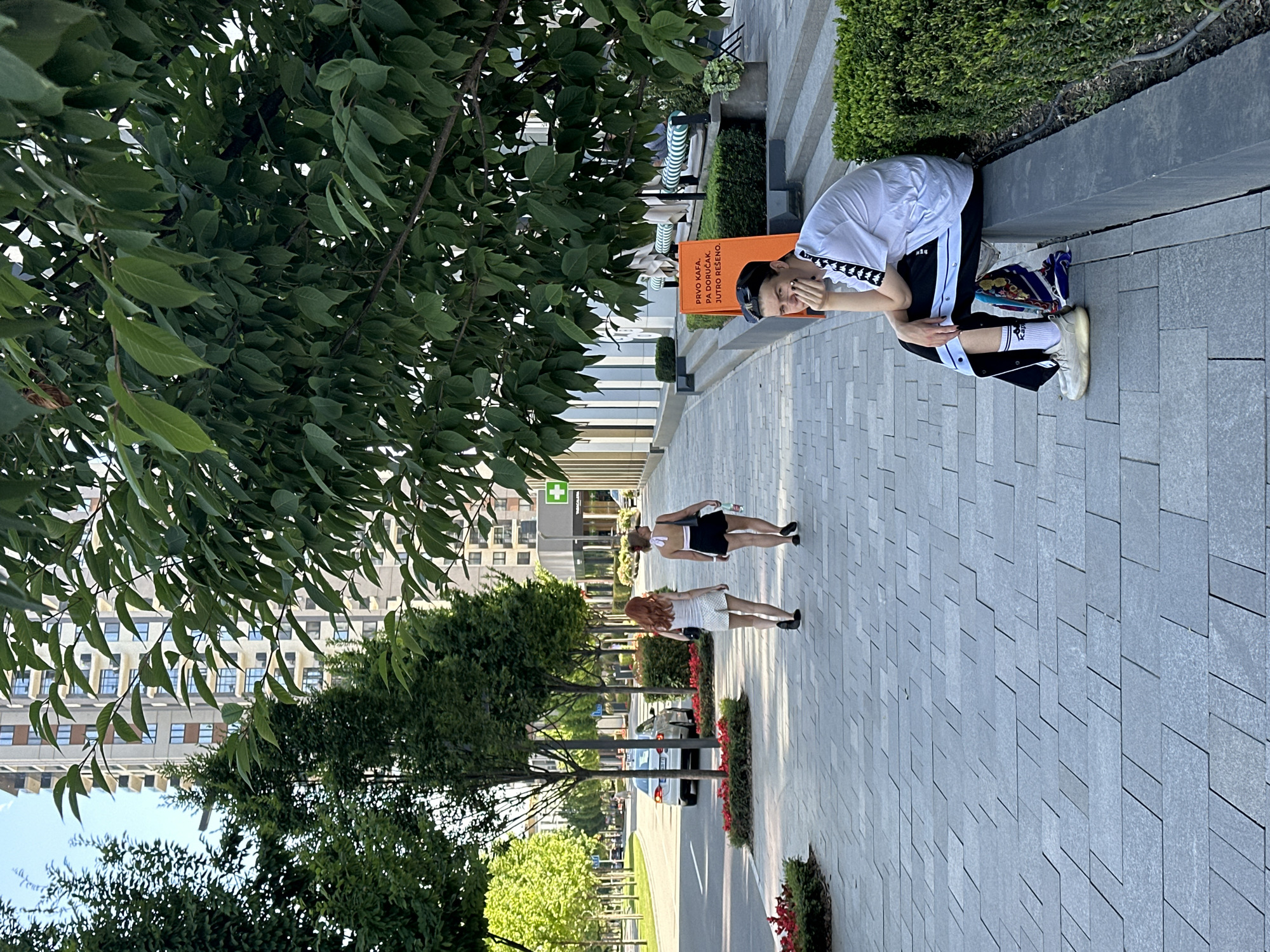 The image depicts a sunny day on a modern, tree-lined pedestrian walkway in Belgrade, Serbia. In the foreground, a young person with short dark hair and sunglasses perched on their head sits on a low stone wall, resting their chin on their hand. They are wearing a white t-shirt with black detailing on the sleeves, black track pants with white stripes and snap buttons, white socks, and light-colored sneakers. A colorful scarf is draped over their lap.
Further down the walkway, two young women are walking away from the camera. The woman on the left has long red hair and is wearing a white tank top and white shorts. The woman on the right has dark hair tied up and is wearing a black halter-neck top and black shorts, carrying a water bottle.
To the left of the walkway is a street with a silver car and trees lining the median. In the background, a tall, modern apartment building with numerous balconies and windows rises into the blue sky. To the right of the walkway, an orange sign with Cyrillic text reading "PRVO KAFA, PA DORUCAK, JUTRO RESENO." is visible, suggesting a cafe or restaurant. Green hedges and benches are also present in this area. The ground is paved with grey rectangular tiles. The overall scene conveys a sense of urban life on a pleasant day.