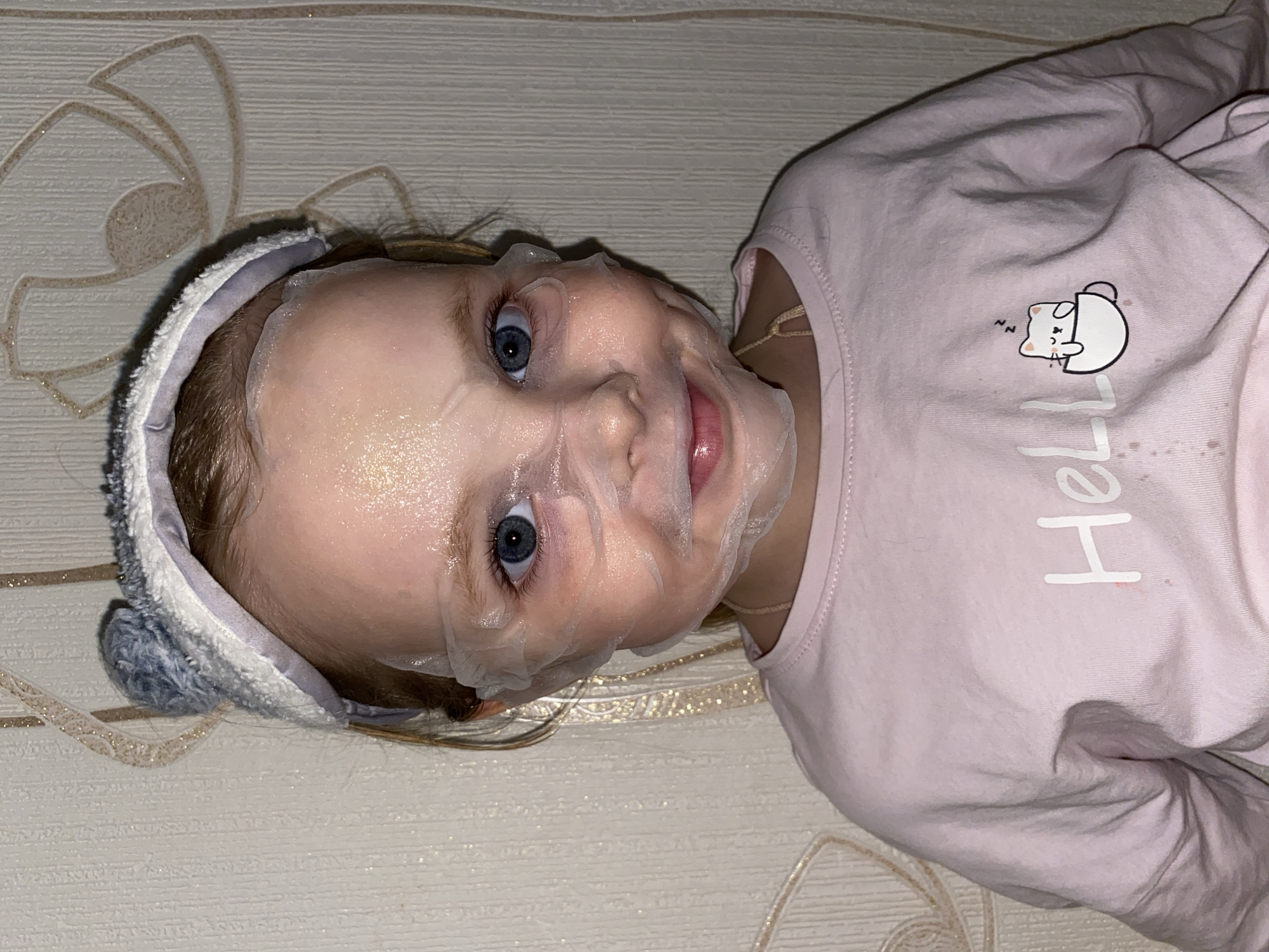 A young child, likely a toddler or preschooler, is depicted lying down and looking directly at the camera with a slight smile. Their face is covered by a translucent, moist sheet mask, which has cutouts for their blue eyes, nose, and mouth. The mask appears shiny from reflected light.
The child wears a grey and white fuzzy headband, possibly styled with animal ears, on their head. They are dressed in a light pink, long-sleeved shirt. A thin, light-colored chain or necklace is visible around their neck. The shirt features a cartoon graphic of a white cat sleeping inside a teacup, with "zzz" indicated above its head. Below this graphic, part of a word, "He," is visible in white text, likely part of "Hello" or a similar word. Some small brown smudges or stains are also present on the shirt.
The child is resting on a light-colored surface, which seems to be wallpaper or bedding, patterned with subtle, light brown or gold floral or swirl designs. The lighting is artificial and indoors. The image is rotated 90 degrees counter-clockwise, with the child's head towards the left side of the frame. The scene captures a playful and candid moment.