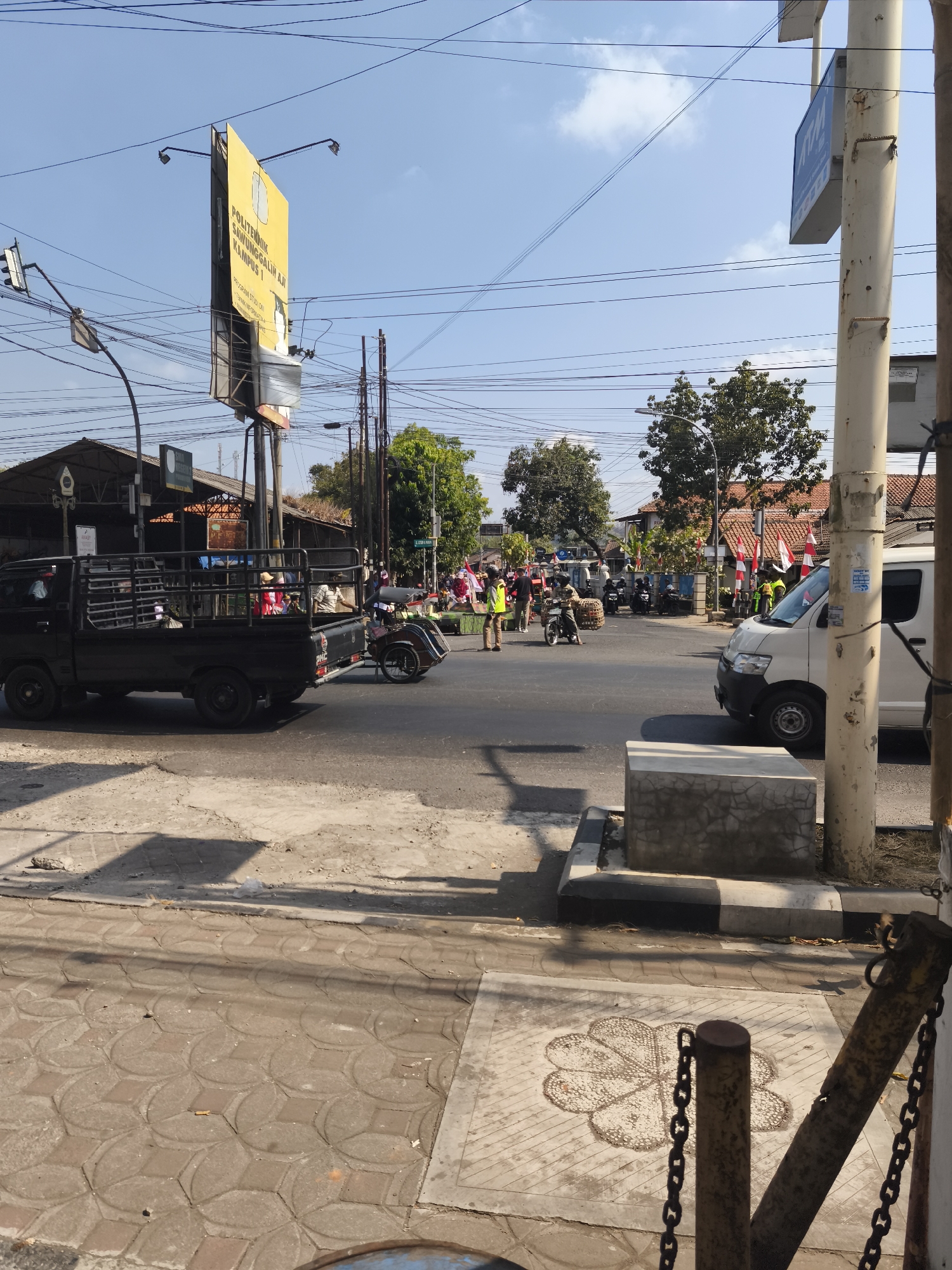 This image captures a street scene in Kutoarjo, Indonesia, possibly near a market or transportation hub, under a clear blue sky. In the foreground, a textured sidewalk with a floral pattern is visible, bordered by a concrete barrier and chain fences.
Dominating the mid-ground is a busy intersection with several vehicles and pedestrians. A black pickup truck with a cage on its bed is parked on the left, facing away from the camera. To its right, a white van is parked on the road. Several individuals in yellow vests, likely vendors or workers, are present near the intersection. Motorcycles, some carrying goods in baskets, are also visible. Red and white flags are strung along the buildings in the background, suggesting a local celebration or event.
A prominent yellow billboard with Indonesian text, likely advertising "POLITEKNIK SAWUNGGALIH AJI KAMPUS 1," stands tall on the left side of the image. Power lines crisscross the sky above, and trees with green foliage are scattered throughout the scene, indicating an urban or semi-urban environment. The overall impression is of an active street with commercial and everyday activities taking place.