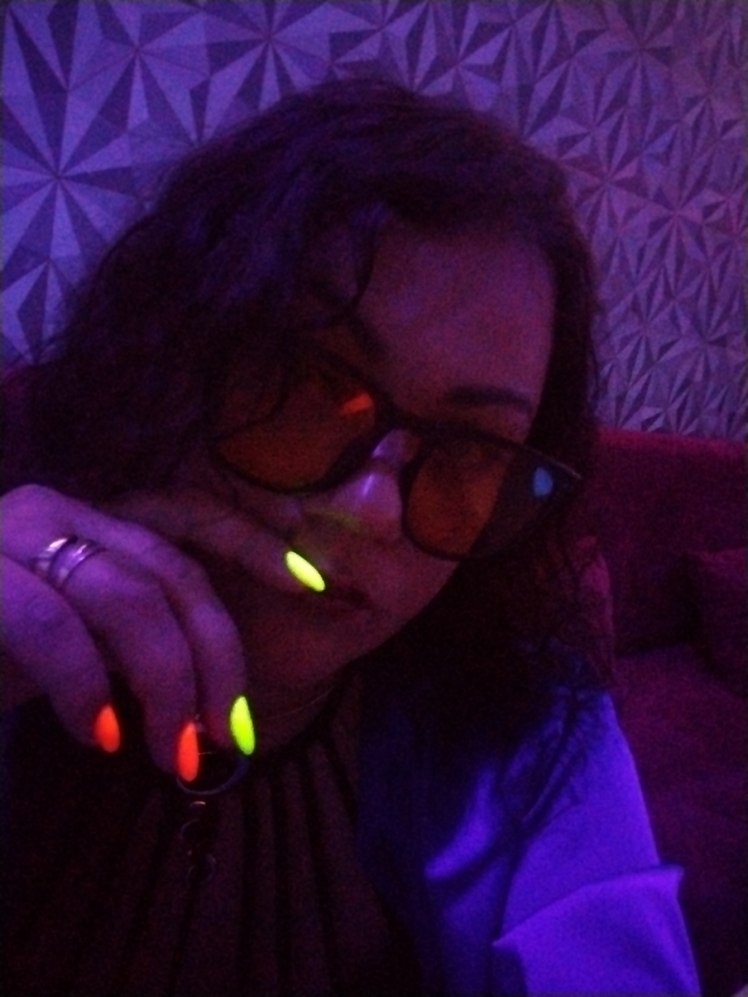 A close-up image depicts a person, likely female, under blacklight illumination in an indoor setting. The individual has dark, curly hair and is wearing large, dark orange-tinted sunglasses that cover their eyes. A silver-toned ring is visible on their left ring finger. Their fingernails are painted with fluorescent polish, with three visible nails glowing bright orange and two glowing neon yellow-green. A dark necklace or pendant is partially visible below the chin.
The background features a wall with a distinctive purple geometric pattern, resembling faceted or tessellated shapes, illuminated by the blacklight. To the right, a section of a dark red or maroon sofa or cushioned seating is visible. The overall scene suggests a dimly lit interior, possibly an entertainment venue, with a playful and vibrant atmosphere created by the UV light and glowing accessories. The activity appears to be a casual pose for a photograph, highlighting the glowing nails. The time of day is clearly nighttime or an equivalent indoor condition. No visible text is present in the image.