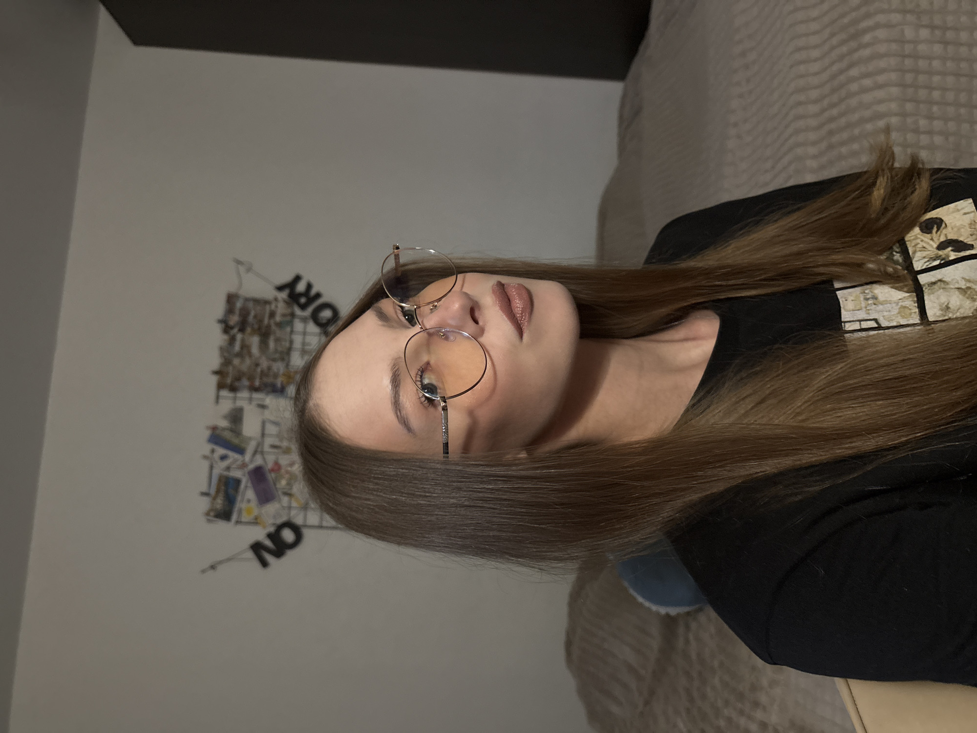 This is a vertical, medium-shot, indoor photograph of a young woman wearing glasses and a black t-shirt. The woman is positioned in the center of the frame, with her head tilted back and her eyes looking upwards. Her hair is long and brown, falling over her shoulders. She is wearing round, gold-rimmed glasses with tinted lenses. Her lips are slightly parted, and she appears to be wearing lipstick.
The background is dimly lit and consists of a wall with some decorative items and text. On the wall, there is a grid-like structure with several small pictures or cards attached to it. Visible text on the wall includes the word "STORY" spelled out vertically, with some letters appearing to be cut off. Below the grid, the word "ON" is visible, written in large, black block letters. The lighting in the room appears to be artificial, suggesting it might be nighttime or a dimly lit interior. The overall mood of the photograph is somewhat mysterious due to the lighting and the woman's expression. The location context is Perm, Russia.