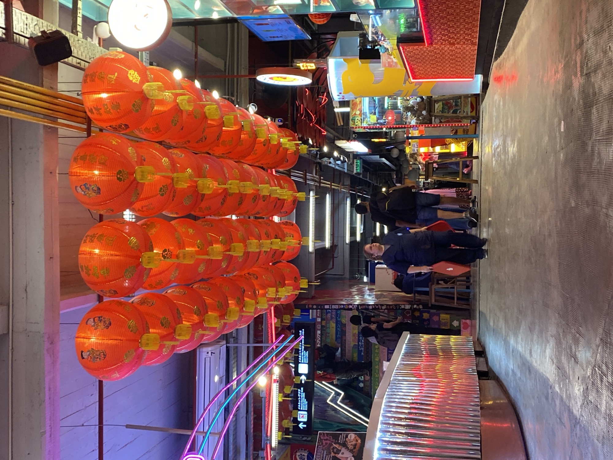Here is a description of the image:
The image shows an indoor scene with a slightly blurred view of what appears to be a bustling marketplace or food court. In the upper-left quadrant, there are rows of illuminated orange lanterns with yellow adornments hanging overhead. These lanterns seem to be the primary focal point, casting a warm glow.
The background reveals a variety of illuminated signs, advertisements, and decorative elements, including what appears to be neon lights and assorted commercial displays. Several people can be seen walking through the marketplace, most of whom are facing away from the camera. A man in a blue uniform is walking through the scene. There is a ribbed metal structure in the foreground.
There are signs with words and symbols in Japanese. The language and architecture suggest a commercial or entertainment district with Asian influence. There are indications that this scene is likely not located in Buenos Aires, Argentina.