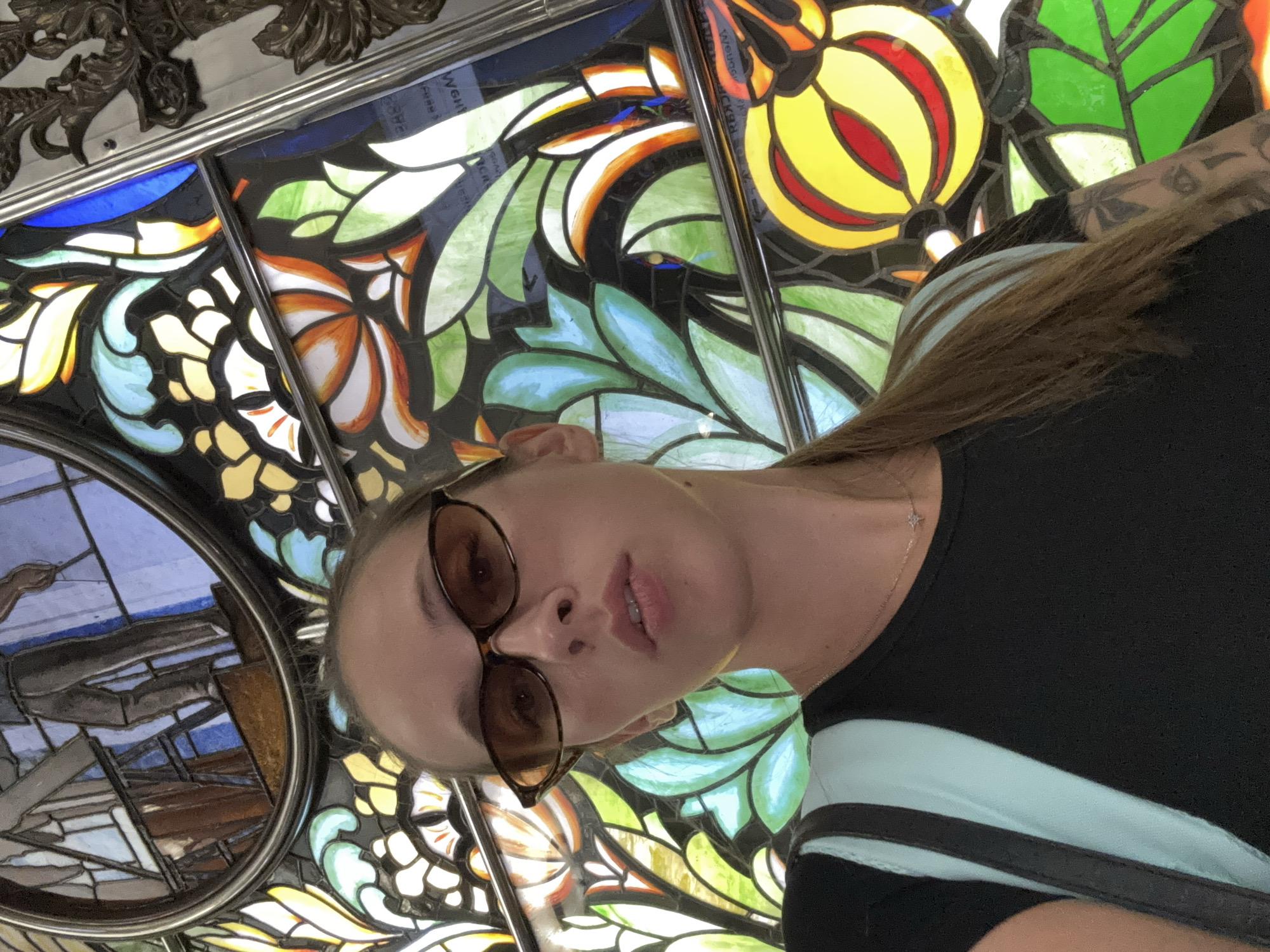 A low-angle, medium shot depicts a woman in front of a stained-glass window. The woman has light skin, long brown hair, and is wearing sunglasses. She wears a black shirt with a light blue and white striped sleeve and a thin necklace. The background is a stained-glass window with a complex design of flowers and leaves in various colors, including blue, green, yellow, orange, and white. The stained-glass window is likely in the Novoslobodskaya metro station in Moscow, Russia.