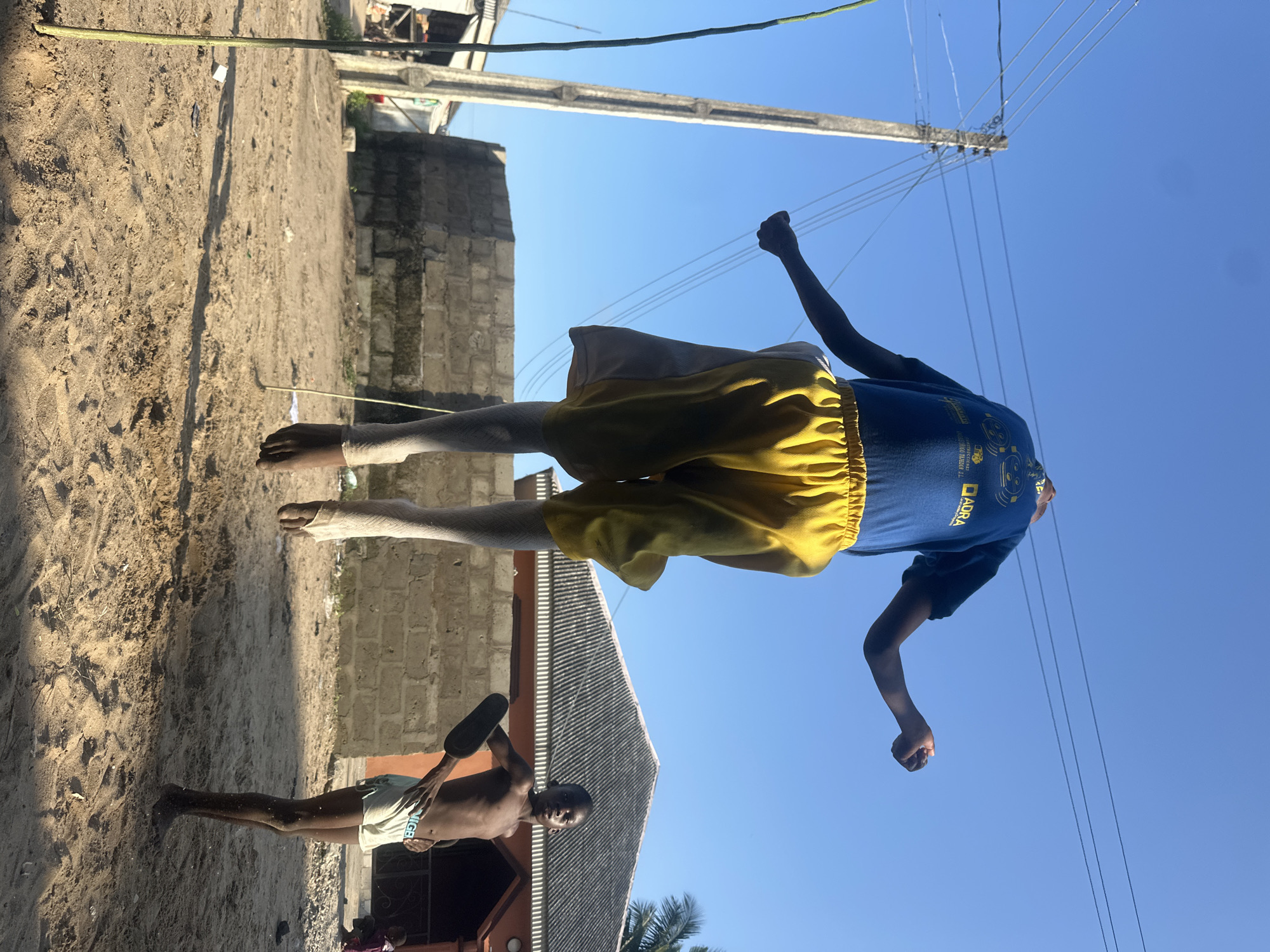 The image depicts two young boys outdoors in what appears to be a rural or semi-urban area in Badagry, Nigeria. One boy, in the foreground and on the left, is standing shirtless and wearing light-colored shorts. He is holding a dark-colored object, possibly a paddle or a flat piece of wood, in his right hand. He is looking towards the camera with a neutral expression.
In the background, a second boy is shown from the waist up, positioned higher in the frame. He is wearing a blue t-shirt and yellow shorts. His left arm is raised and his fist is clenched. He seems to be looking upwards or outwards.
The setting is characterized by sandy ground with some scattered debris, and a brick building with a corrugated roof. A tall wooden pole, likely supporting electrical wires, is visible in the background, stretching up into a clear blue sky. The sun is casting shadows, suggesting it is either morning or late afternoon. The overall atmosphere appears to be one of everyday activity in a warm climate.
Visible text on the blue t-shirt includes "ADRA" in a yellow box, and some other text that is partially obscured or too small to read clearly.