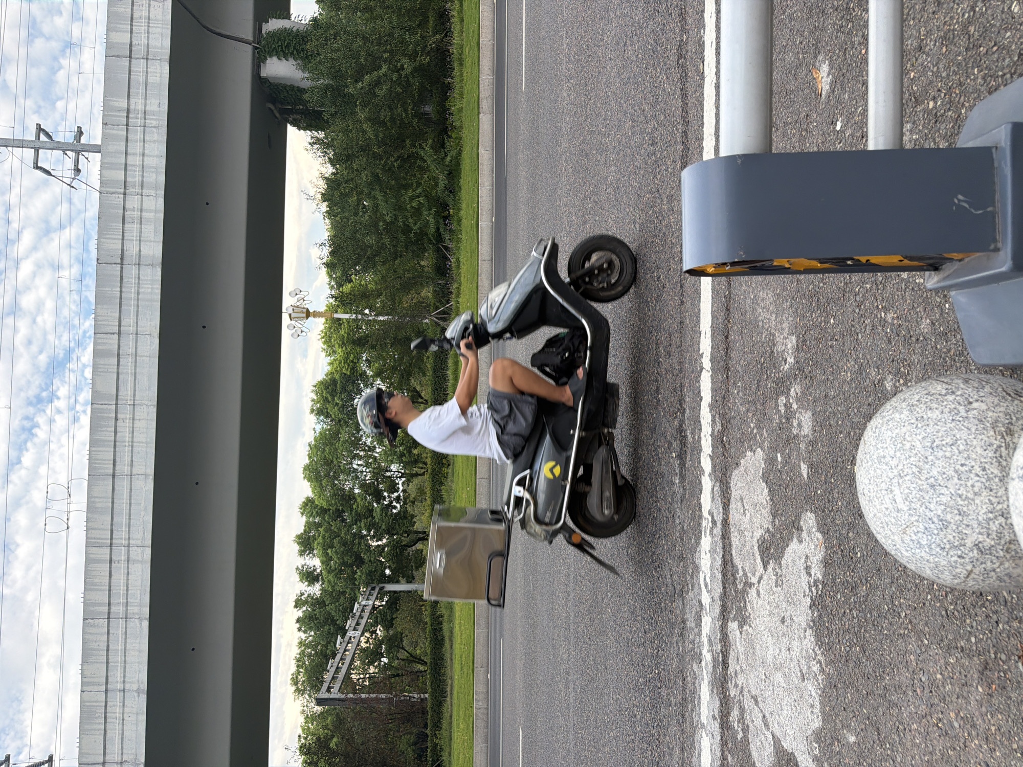 In Jiangjin, China, a person wearing a helmet and a white t-shirt rides a dark gray electric scooter on a paved road. The scooter has a silver cargo box mounted on the back. The rider is wearing dark shorts and sandals. The road is lined with grass and trees, and an elevated highway structure is visible in the background. The sky is partly cloudy. The scene appears to be on a street with traffic.