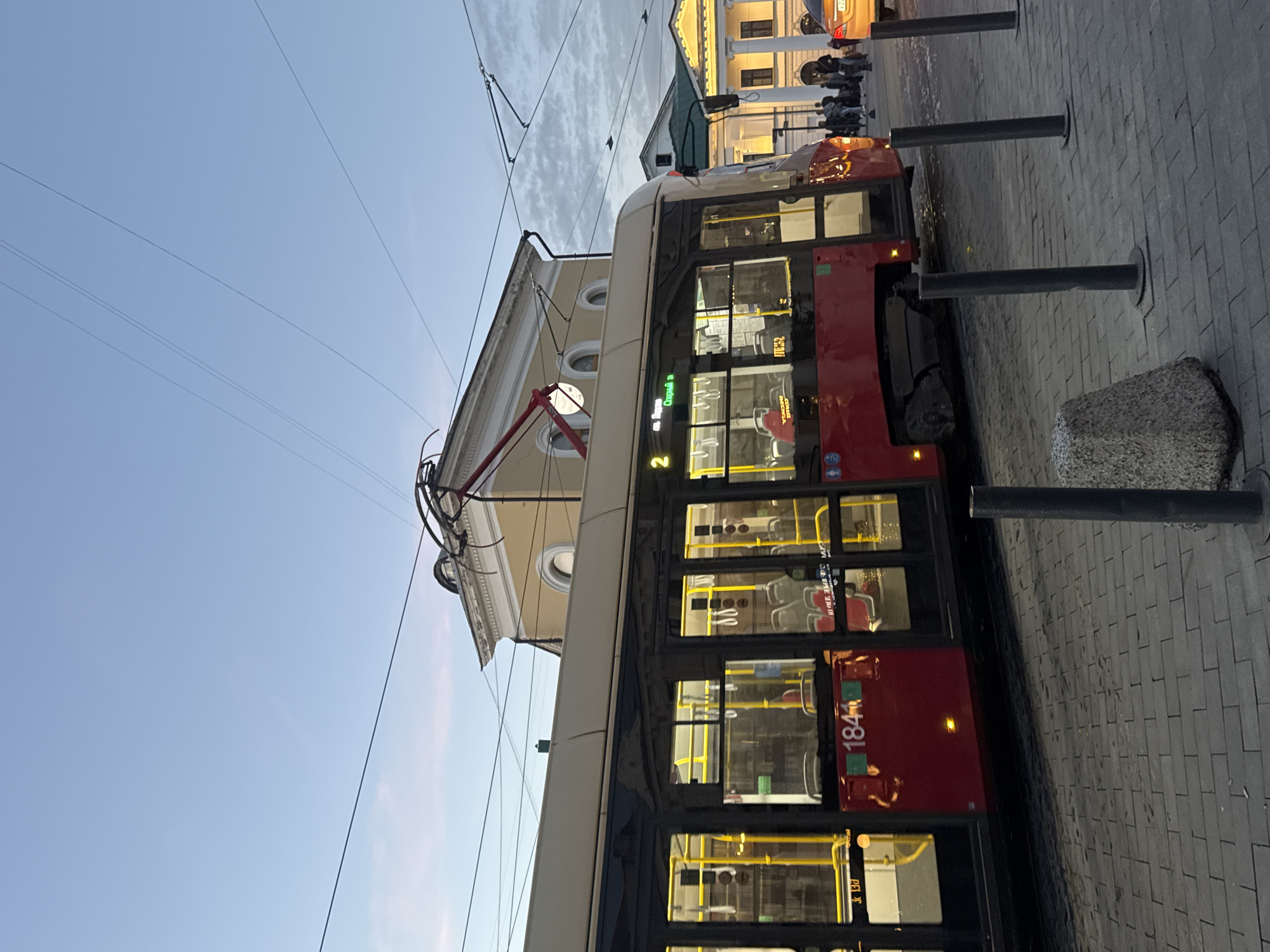 The image depicts a street scene in Nizhny Novgorod, Russia, at dusk. A red and beige tram is the central subject, positioned on the right side of the frame. The tram's windows are illuminated from within, revealing rows of empty red seats and yellow vertical poles. On the exterior of the tram, the number "1844" is visible in green digits.
Above the tram, overhead wires and power lines crisscross against a pale blue sky with wispy clouds. Behind the tram, a pale yellow building with arched windows and ornate architectural details is visible. To the right of this building, another white building with columns and arched windows is illuminated, suggesting it is a public or historical structure.
On the sidewalk in front of the buildings and to the right of the tram, a small group of people is gathered, their faces indistinct. A yellow car is parked on the street to the far right. The ground is paved with cobblestones. Several dark gray bollards are planted in a row along the edge of the sidewalk, and a large, rough-hewn stone object is positioned on the left side of the foreground. The overall impression is of a public space in the city during the transition from day to night.