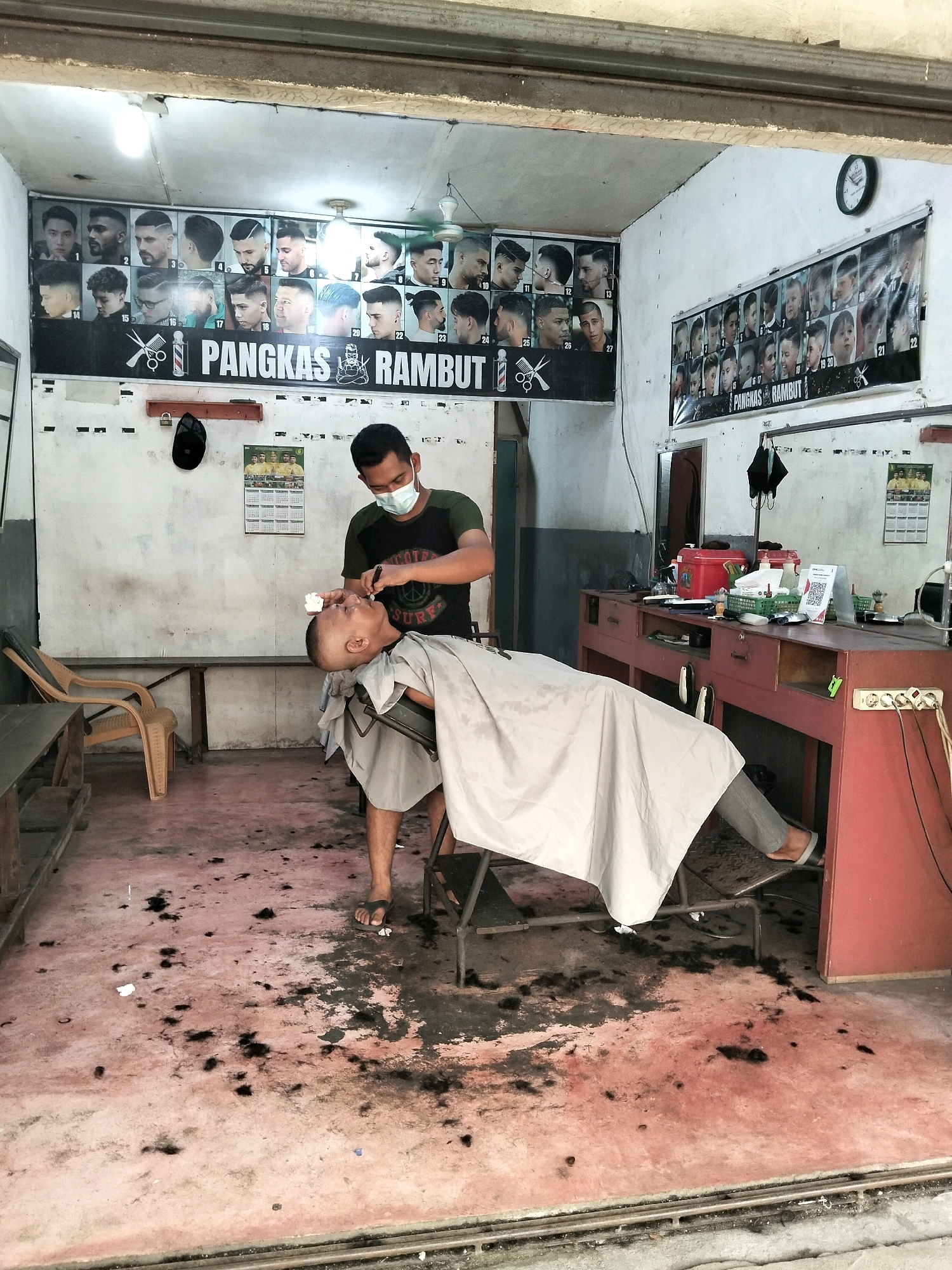 In a modest barbershop in Arang Limbung, Indonesia, a barber wearing a mask and a dark t-shirt is carefully shaving the head of a customer who is reclined in a chair and draped with a light-colored cape. The floor of the barbershop is littered with clippings of dark hair, indicating recent haircuts. Behind the barber, a row of framed images displays various hairstyles. A red barber's chair with metal armrests is visible. A barber's station with drawers and shelves is situated to the right, holding various barber tools and products. The walls are painted white, and a clock is mounted on the upper right wall. A chair, possibly a waiting chair, is positioned to the left. The overall impression is of a functional, working barbershop.