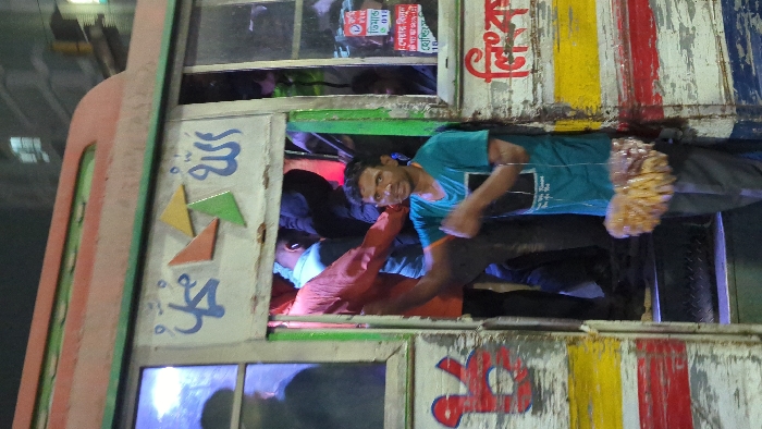 A crowded bus with passengers hanging out of windows. The exterior of the bus is decorated with colorful paint and what appears to be Arabic script. One man in a blue shirt is visible, leaning out of a window with a slight smile. The scene suggests a public transportation scenario, possibly in a densely populated area.