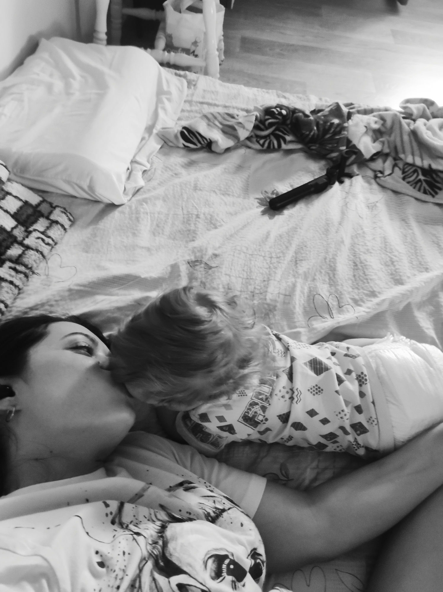 A black and white photograph shows a woman and child lying on a bed. The woman is lying on her back, with her head tilted to the right and her eyes closed. She appears to be kissing the child's head. The child is lying face down on the woman's chest, with its back to the camera. The child is wearing a patterned top and a diaper. The bed is covered in a patterned sheet and blanket. A pillow is visible at the top left of the frame. A black object resembling a remote control or hairbrush is on the bed near the top right corner of the frame. A small heart-shaped design is visible on the bedspread near the top left corner of the image. The location appears to be a residential interior.