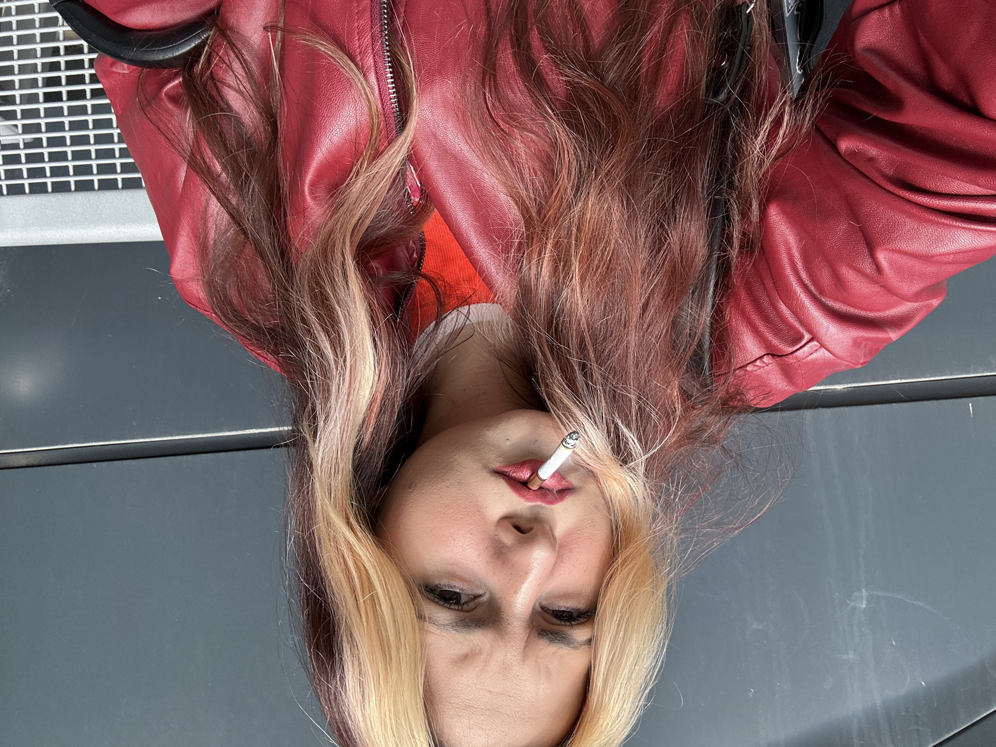 A fair-skinned woman with blond and auburn hair is seen from the chest up, with a cigarette in her mouth. She wears a red leather jacket. The background is a combination of grey and silver metal. The image does not provide enough information to determine a location in Sofia, Bulgaria.