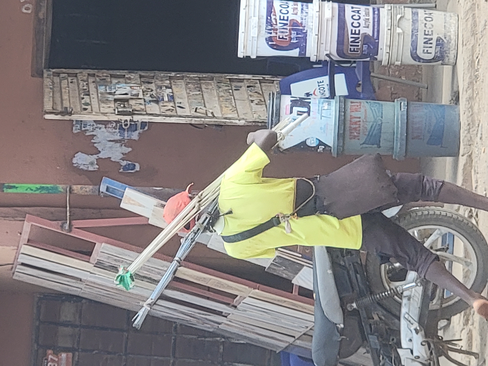 In Maiburiji, Nigeria, an individual wearing a yellow long-sleeved shirt and dark pants is positioned on a white motorcycle. The motorcycle is loaded with bundles of wooden planks and silver metal rods tied together with green material. The surrounding environment includes paint buckets, and a building with a weathered facade. The person is wearing a red cap and a black strap across the chest.