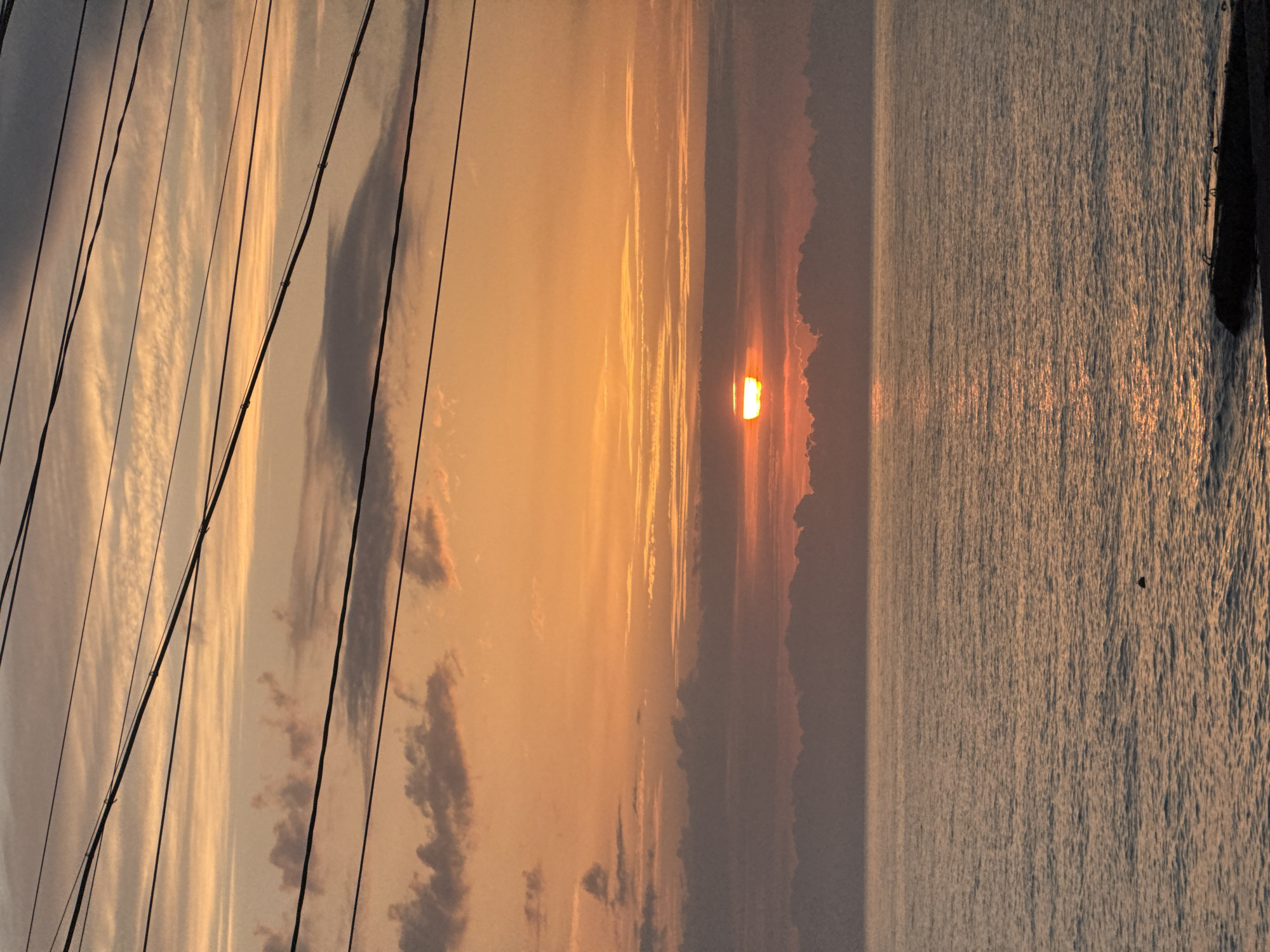 The image depicts a sunset over water, possibly a sea or large lake, with the view partially obscured by lines or rigging of a boat. The sky transitions from a pale blue on the left to a golden orange towards the center where the sun is setting. Wisps of clouds are scattered throughout the sky. The water reflects the golden light of the sunset. The location is not discernible from the image alone, so I cannot specify it is in Sochi, Russia.