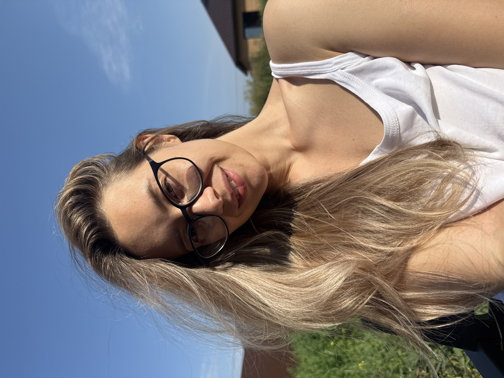 A woman with blonde hair and glasses is taking a selfie outdoors. She is wearing a white tank top. In the background, there is green grass, a blue sky, and houses. The image appears to have been taken on a sunny day. The location is in the city of Chaikovsky, Russia.