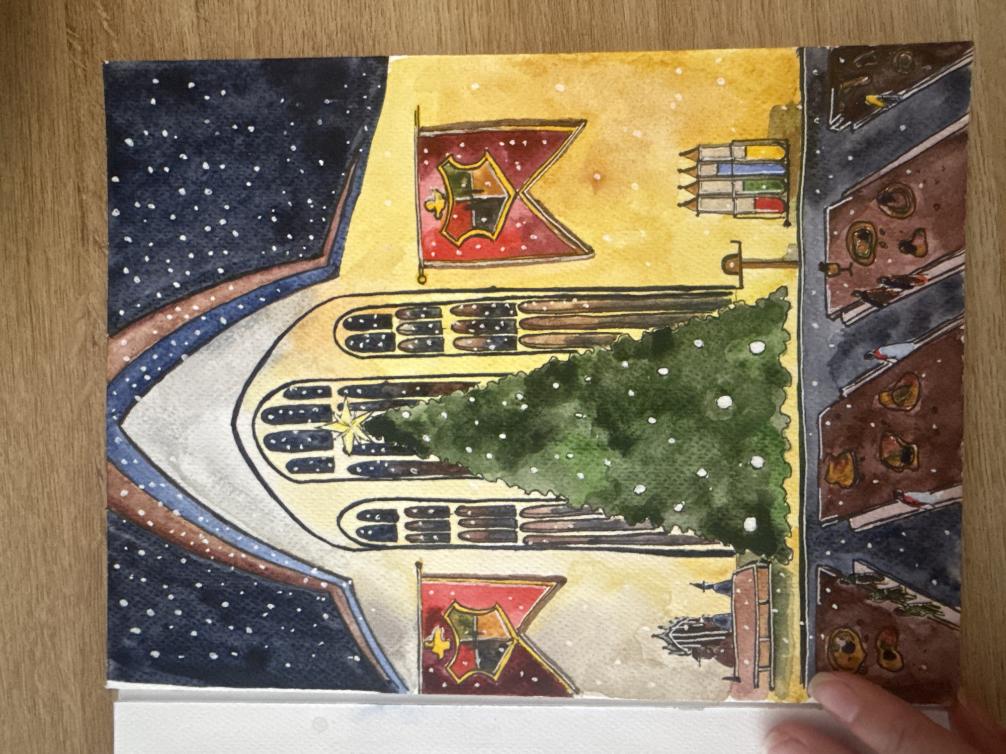 A watercolor painting depicts a festive scene inside a grand hall, likely a Great Hall, illuminated by warm yellow light and a starry night sky visible through arched windows. A large, decorated Christmas tree stands in the center, adorned with white ornaments and topped with a golden star. Red and gold banners bearing a crest are hung on either side of the hall, suggesting a school or house affiliation. In the foreground, long tables are set for a meal, with food and drinks laid out. Small figures of people can be seen seated at the tables. A rack of colorful vials sits on a shelf to the right. The painting's style is whimsical and illustrative, with a light dusting of snow falling outside. There is no specific visual information within the painting that places it in Wrocław, Poland; it is a general depiction of a fantasy or historical setting.