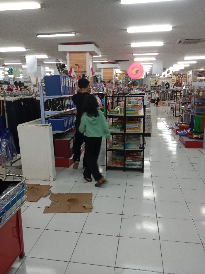 The image depicts an indoor retail environment located in Langenharjo, Indonesia. The floor is composed of white tiles, some exhibiting hairline cracks, with three irregular pieces of brown cardboard placed in the immediate foreground; one piece shows a distinct dark stain. The white ceiling features a grid of evenly spaced fluorescent tube lights and multiple air conditioning vents.
In the mid-ground, two individuals are walking away from the camera. The person on the left wears a dark top. The person on the right, slightly ahead, wears a light green hooded top and dark trousers, with their feet blurred in motion. They are positioned to the right of a black metal shelving unit containing multiple shelves filled with books and various merchandise. To their left, a tall, light blue shelving unit holds several blue ring binders, adjacent to a white rectangular object. Further left, clothing racks display dark garments, and a blue plastic bag is visible.
The background shows additional retail display units stocked with a variety of products. A large, pink, donut-shaped inflatable object is suspended from the ceiling in the central background. A structural pillar with light wooden paneling is adorned with two small flags resembling the Canadian national flag. A third individual, partially obscured, is visible walking in the far right background. A red-framed metal shopping basket on wheels is partially visible in the lower-left foreground.