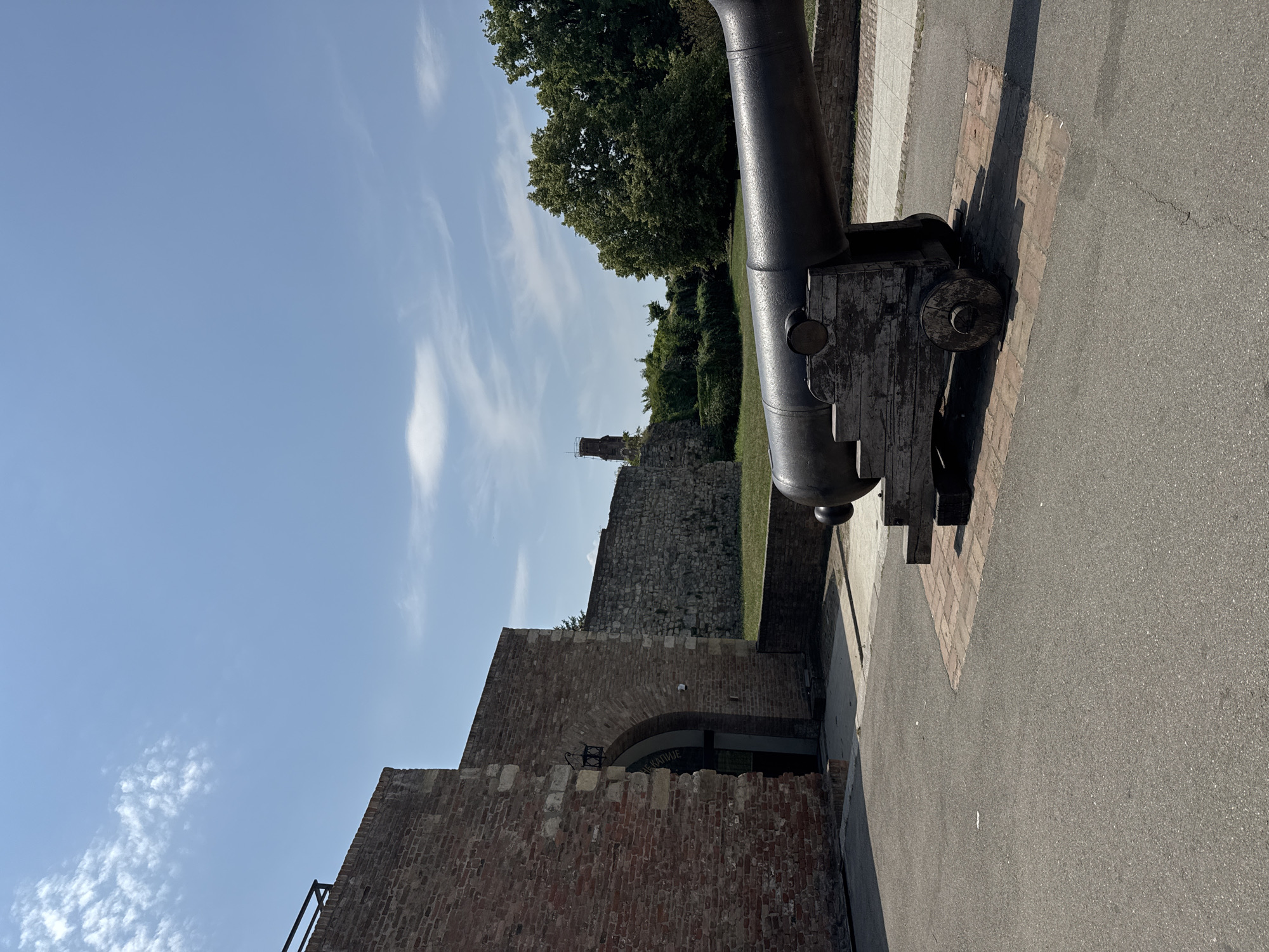 The image shows a historical scene at the Belgrade Fortress in Belgrade, Serbia, on a clear, sunny day. In the foreground, a large, dark cannon with a wooden carriage sits on a brick-laid surface. The cannon is positioned on the right side of the frame, with its barrel pointing towards the left. Behind the cannon, a section of the fortress wall, constructed from red brick, is visible on the left. An arched entryway is present in this brick wall, with a black lamp fixture above it. Further back, another stone fortress wall stretches across the midground, with lush green grass and trees behind it. In the distance, a tall, reddish-brown tower or structure is visible against the bright blue sky, which has scattered white clouds. The ground is paved with asphalt.