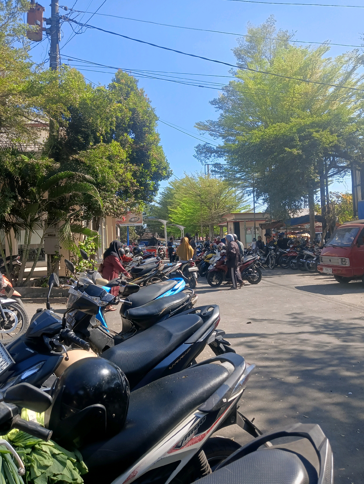 The image depicts a street scene in Sokaraja, Indonesia, likely a public area or marketplace. A large number of parked motorcycles are visible, dominating the foreground and extending into the mid-ground. The motorcycles are of various models and colors. Several people are present, some walking, some appearing to interact with the motorcycles, and others standing around. They are dressed in casual clothing, with some women wearing hijabs. In the background, buildings are visible, along with trees providing shade. A red van is parked on the right side of the image. The sky is clear and blue, suggesting a sunny day. The overall impression is of a busy, functional urban environment.
