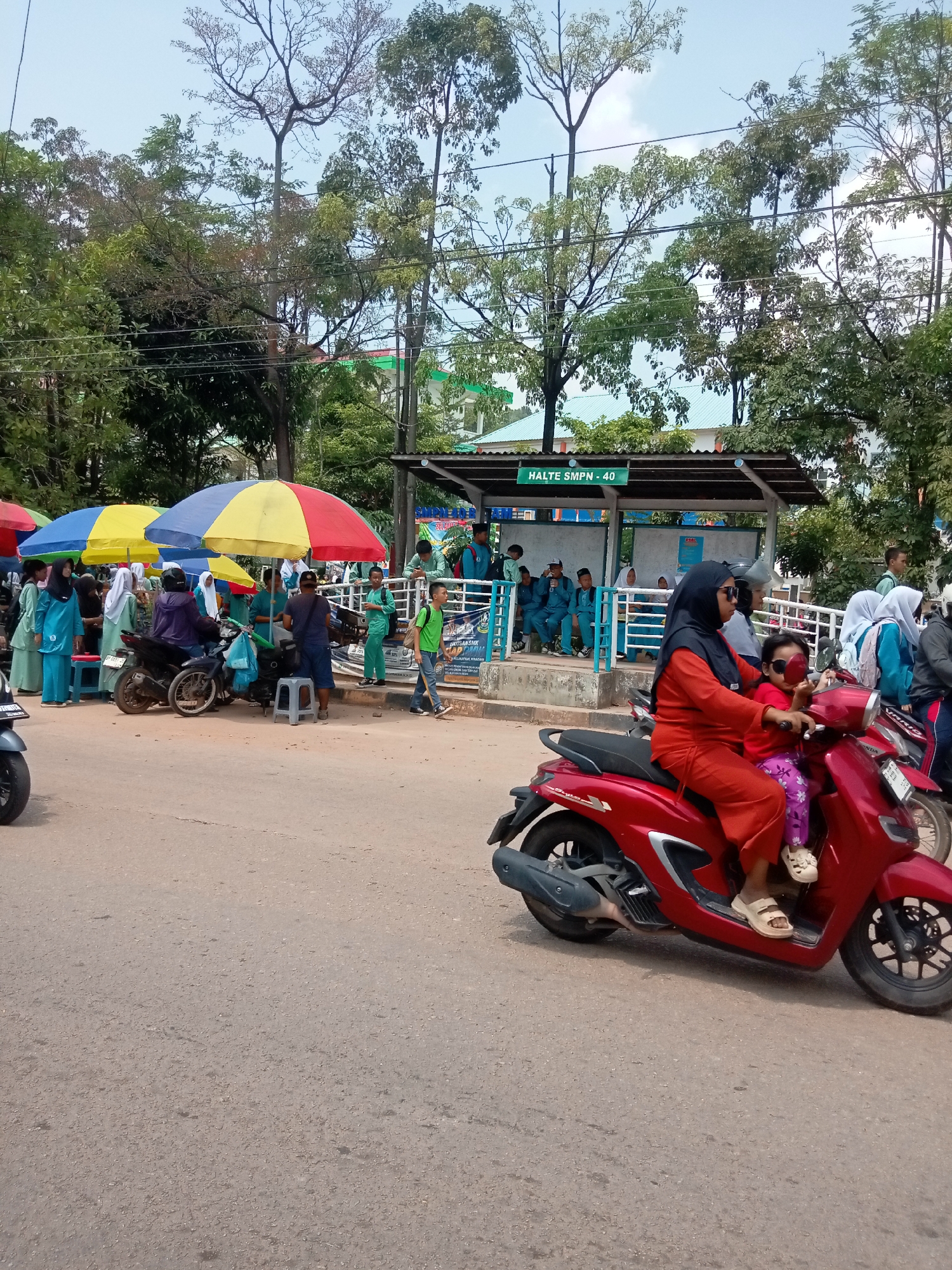 A busy street scene unfolds near Halte SMPN 40 in Batam, Indonesia, under a bright sky with scattered clouds. In the foreground, a red scooter carries a woman wearing an orange hijab and sunglasses, with a young child in a pink shirt and purple pants seated in front of her. The child also wears large sunglasses. Both are facing left and appear to be moving away from the viewer.
To the left, a row of colorful umbrellas shields several people. Some are dressed in light blue and white uniforms, possibly students, gathered around a motorbike and several small stools. A person in a dark jacket is seated on the motorbike. Further left, more individuals in similar uniforms are visible.
In the background, a covered bus stop or waiting area labeled "HALTE SMPN - 40" is situated behind a railing. Inside the shelter, several students in blue uniforms are seated. Beyond the shelter, lush green trees and buildings with green roofs are visible. The street itself is paved with asphalt, showing some cracks and texture. The overall impression is a lively public space, likely a school drop-off or pick-up point.