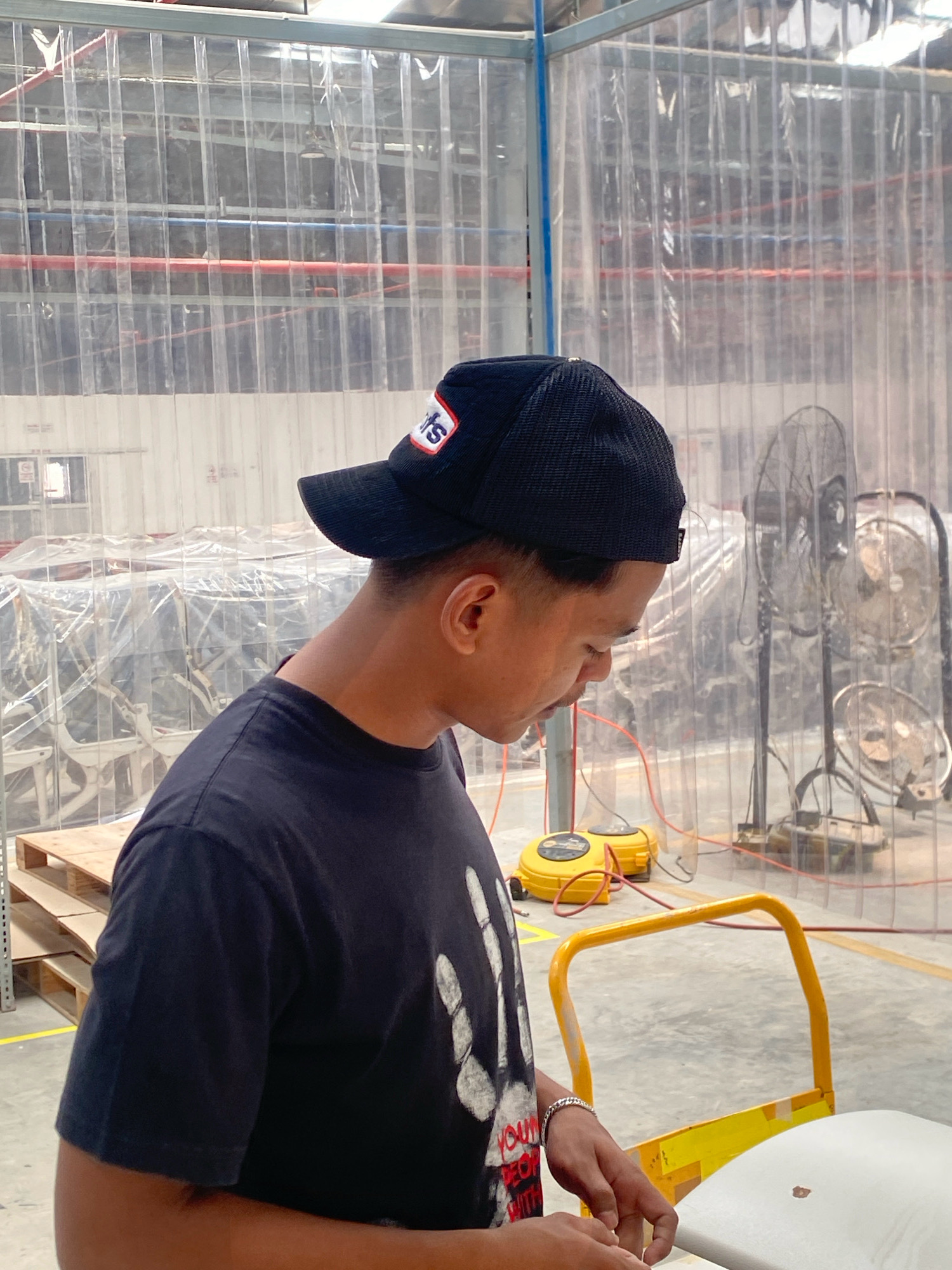 In Batam, Indonesia, an individual wearing a black t-shirt and a black baseball cap with a red and white logo is positioned in what appears to be an industrial or workshop setting. The person is looking downwards, with their hands in front of them, engaged in an activity. The environment is characterized by large, clear plastic sheeting hanging from overhead structures, creating partitions. Behind the sheeting, industrial shelving units with red and blue piping are visible. In the background, several industrial fans are present, along with what seem to be covered materials on pallets. A yellow piece of equipment with two circular components is situated on the floor to the left, connected by wires. A yellow metal bar structure is also in the foreground. The flooring is concrete.