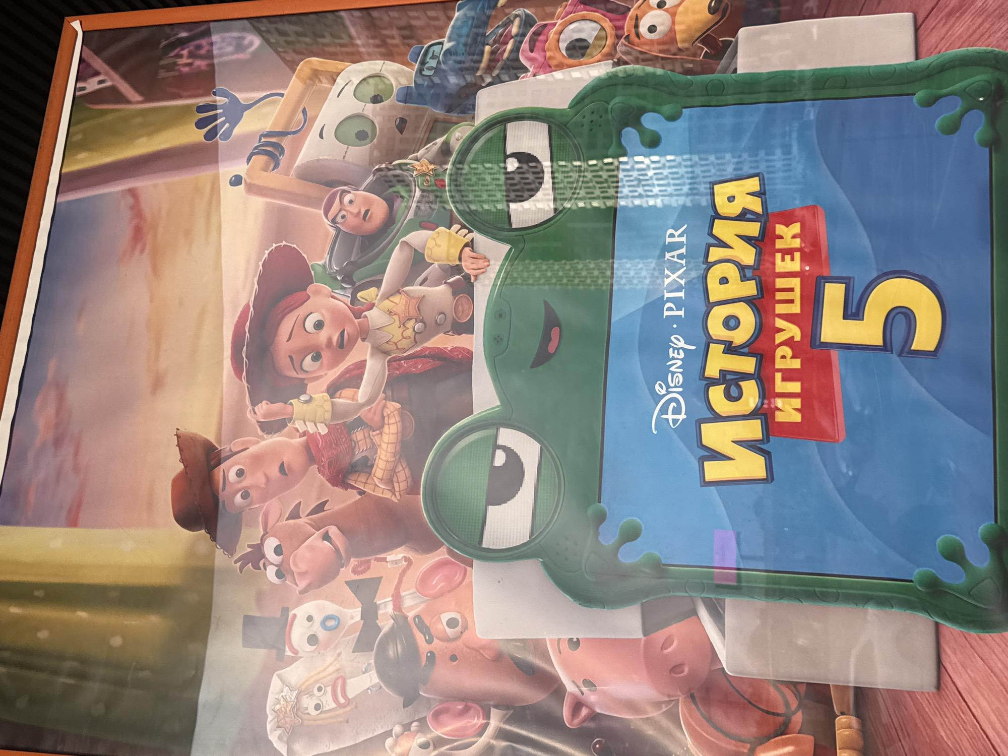 This media file is a photograph of a poster for the movie "Toy Story 5." The poster features a collage of characters from the "Toy Story" franchise, including Woody, Jessie, Buzz Lightyear, and other toys, rendered in their animated style. The characters are positioned in a playful and somewhat chaotic arrangement, with some appearing to interact with each other.
Prominently displayed on the poster is the title, which is in Russian: "ИСТОРИЯ ИГРУШЕК 5" (Istoriya Igrushek 5), meaning "Toy Story 5". The text "Disney · PIXAR" is also visible, identifying the production studios. The background appears to be a stylized representation of a room or playroom, with warm, soft lighting that suggests a domestic setting, possibly indicating daytime. The overall emotion conveyed by the poster is one of fun and adventure, typical of the "Toy Story" series. The poster itself is framed by a reddish-brown border. The image's orientation is rotated, making the text appear vertical. The scene is likely intended for a public display, such as a movie theater or a shop window. The mention of Krasnogorsk, Russia, suggests this poster was photographed in that location.