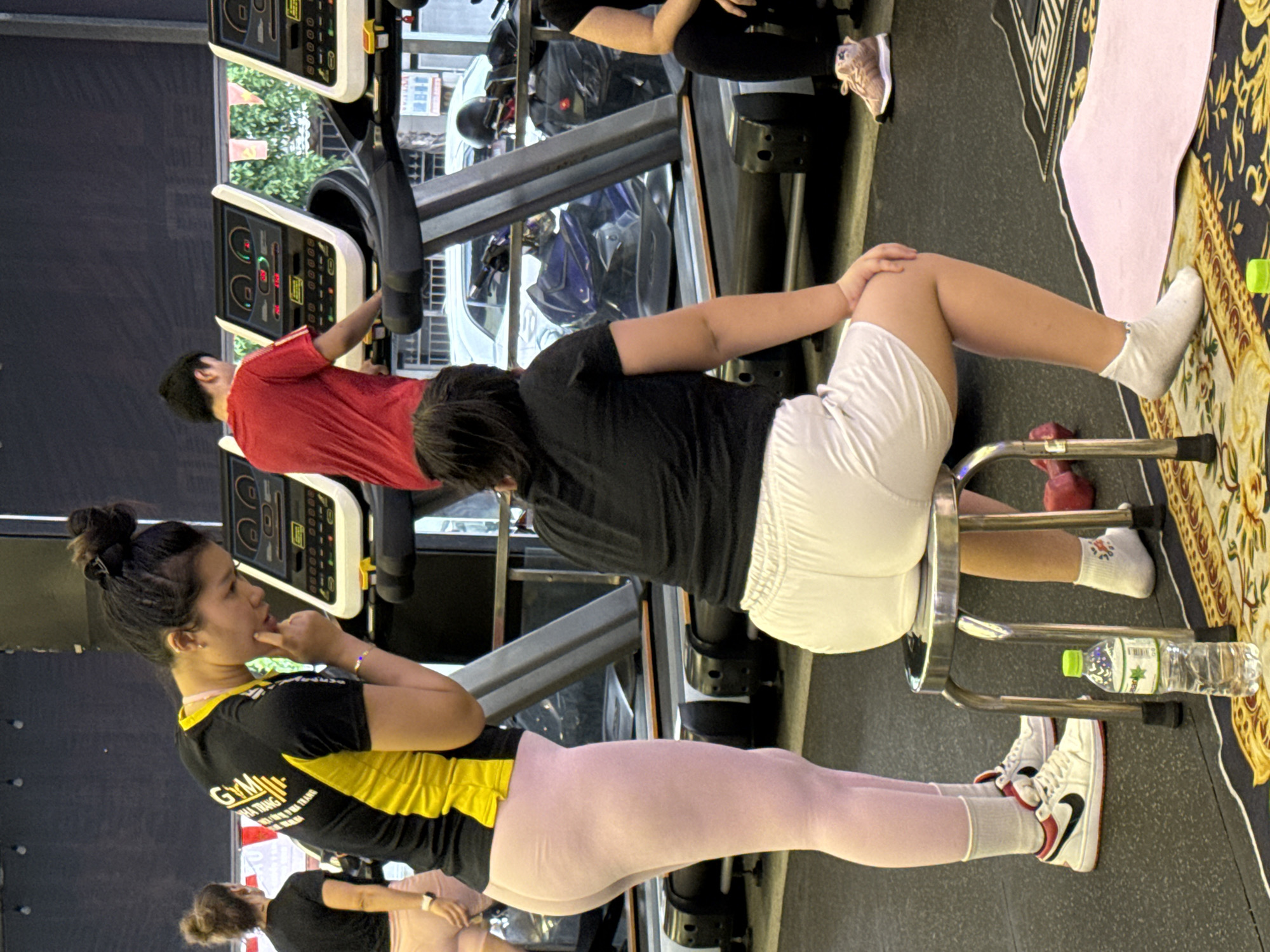 The image depicts an indoor gym environment in Nha Trang, Vietnam, with several individuals engaged in fitness activities. In the foreground, a person is seated on a metallic stool wearing white shorts and socks, with a bottle of water and pink dumbbells visible nearby. To the left, another person stands in pink leggings and white sneakers, wearing a black and yellow gym shirt. In the background, a person in a red shirt uses a treadmill, and another person is partially visible on a treadmill on the far right. The gym appears to be equipped with multiple treadmills and other exercise equipment.