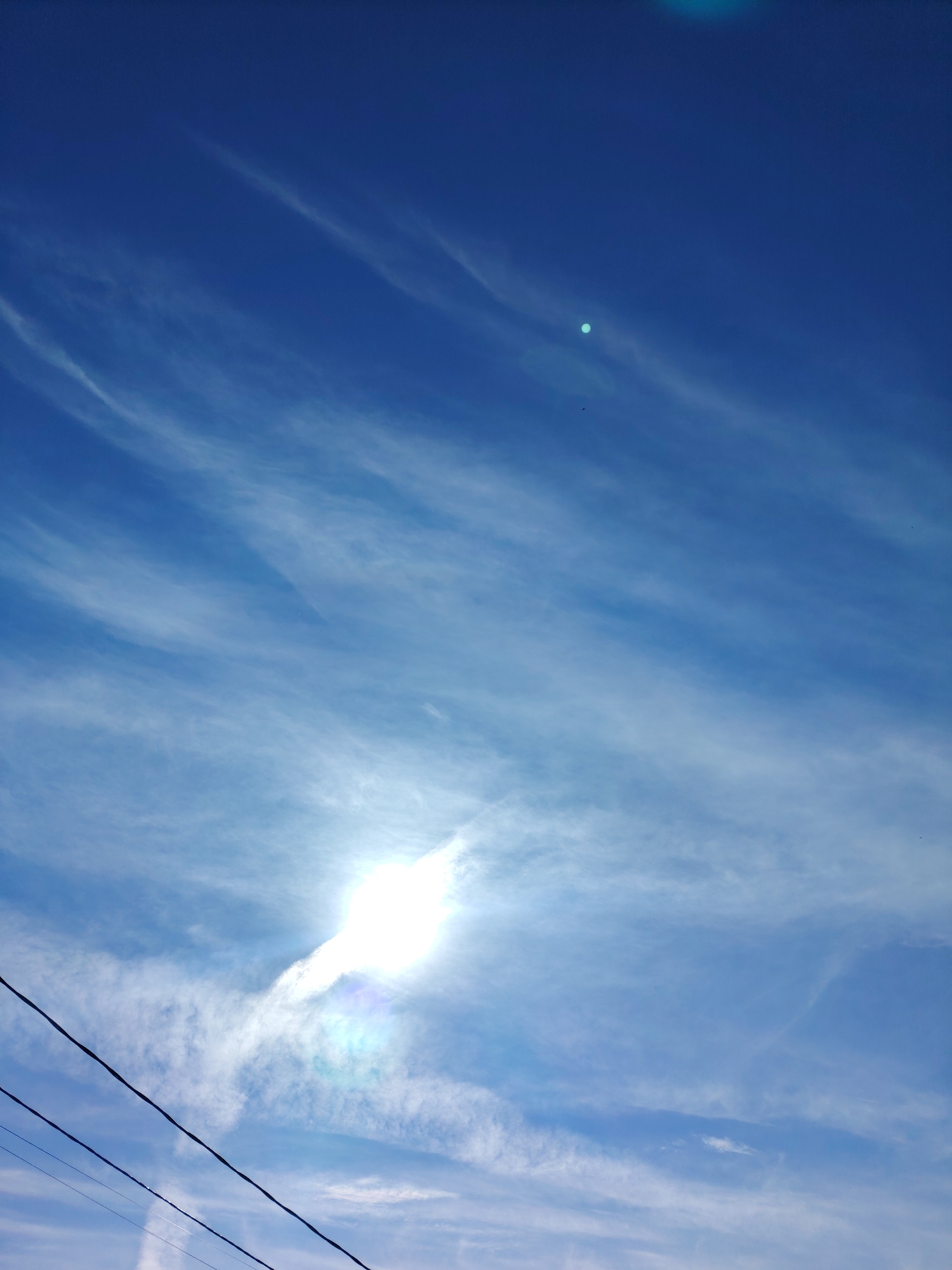 The image captures a bright, expansive blue sky adorned with long, wispy white clouds. The sun is the dominant feature, positioned in the lower-middle of the frame, appearing as an intensely bright, overexposed white light source. This strong illumination creates a distinct lens flare effect, producing shimmering green and blue hues around the sun's perimeter and casting a radiant glow across the surrounding clouds.
Several dark, thin utility wires or power lines cut across the bottom-left portion of the image, running diagonally from left to right. A tiny, dark speck, possibly a bird or insect, is visible in the upper-middle left area of the sky, and another faint speck can be observed in the upper-right corner. The clouds are high-altitude and scattered, forming elongated streaks across the blue expanse, which deepens in color towards the top of the frame. The scene conveys a clear, sunny day in Chaikovsky, Russia, likely during midday due to the high intensity of the sun. No people or visible text are present in the image.