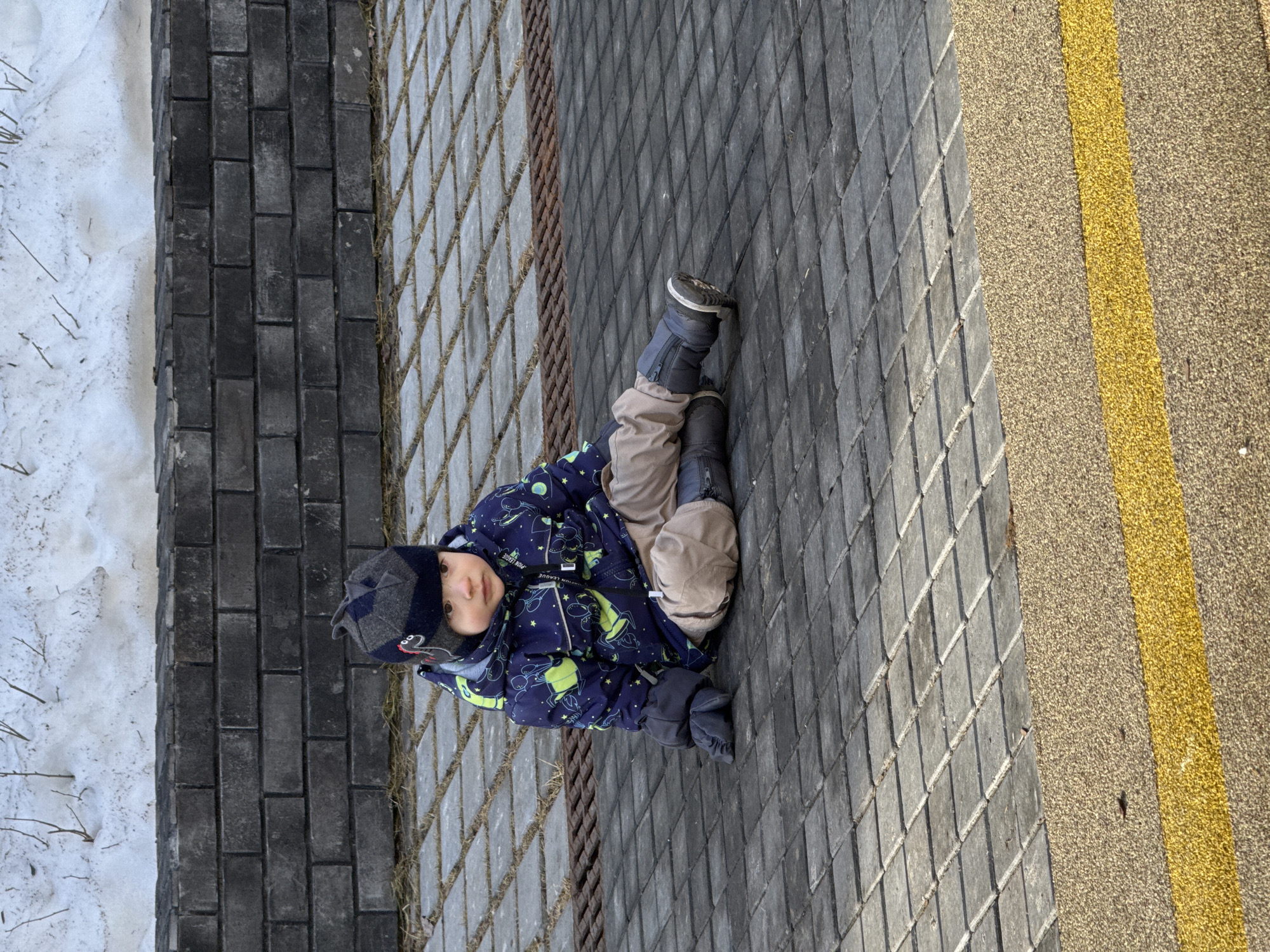 An outdoor scene in Izhevsk, Russia, depicts a young child sitting on a grey paved surface. The child is dressed in a dark grey and blue patterned winter hat with fin-like protrusions, a blue winter jacket featuring green and white patterns, tan trousers, dark grey gloves, and dark blue winter boots with white soles. The child sits with legs extended forward and the left hand resting on the square-patterned pavers. Behind the child, a rusty metal grate is visible, followed by a low wall constructed of dark grey bricks. Beyond the brick wall, a patch of dirty snow with sparse brown vegetation is present. In the immediate foreground, a textured, light brown aggregate surface with a prominent horizontal yellow stripe extends across the bottom of the frame.