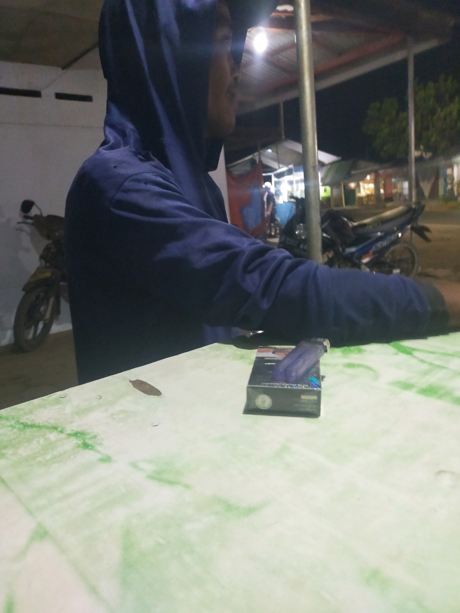 A medium shot captures a person wearing a dark blue hooded jacket, seated at a table at night. The person's face is partially obscured by the hood, but they appear to be looking towards the right side of the frame. A pack of cigarettes and a lighter are placed on the table in front of them. The table surface is a light green color with a marbled pattern. In the background, a motorcycle is visible to the left of the person, and further back, there are streetlights and buildings that suggest an urban or roadside setting, consistent with the location in Gedong Wetan, Indonesia. The lighting is dim, indicating nighttime.