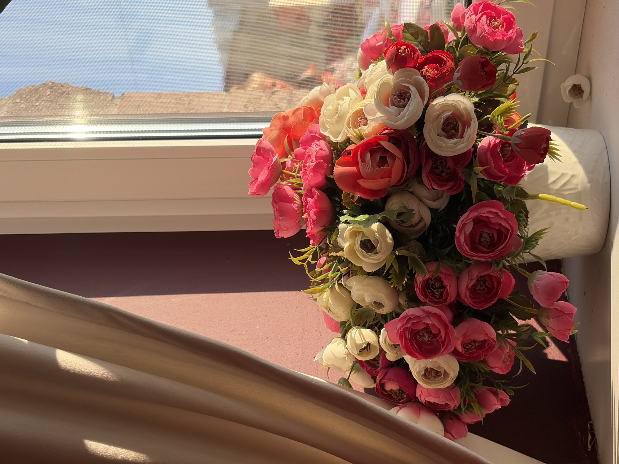 The image depicts a vibrant bouquet of artificial flowers, predominantly in shades of pink, red, and cream, arranged in a white vase. The bouquet is placed on a windowsill, with a view of an outdoor scene through the window. The outdoor scene shows a rooftop and a hillside under a clear blue sky, bathed in bright sunlight, suggesting a sunny day. The light streaming through the window casts shadows on the windowsill and the surrounding area, indicating it is likely daytime, possibly morning or afternoon given the angle of the light.
The windowsill itself is white, and the surface below it appears to be a reddish-brown color. A cream-colored drapery or fabric is visible in the foreground, adding a soft, textured element to the composition and catching the sunlight. The overall impression is one of domestic tranquility, with the artificial flowers adding a touch of color and cheerfulness to the setting. There are no visible people, text, or specific clues to pinpoint an exact location beyond the general context of a window and an outdoor view. The focus is on the still life arrangement of the flowers against the backdrop of the window and the external environment.
