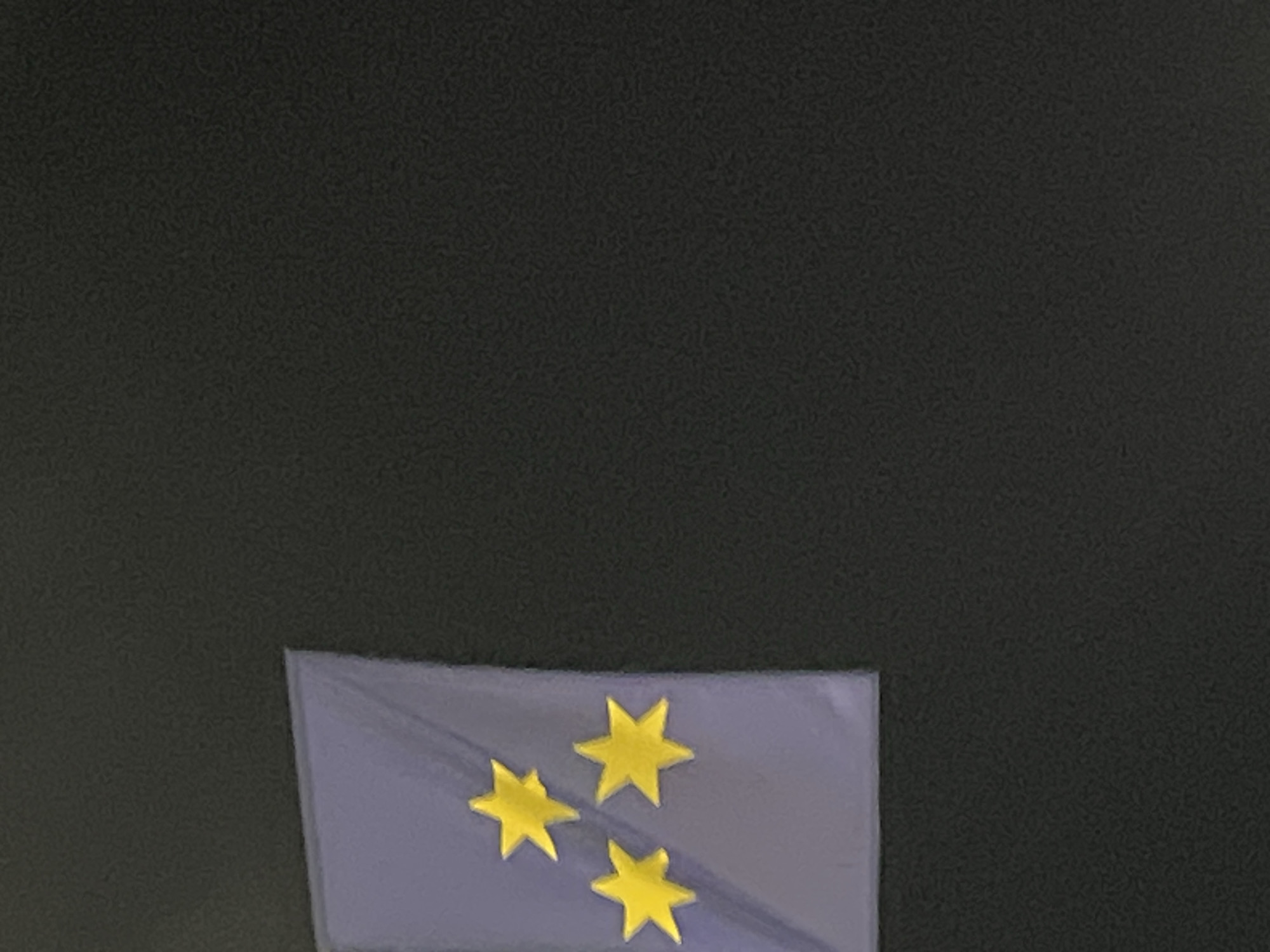 The image shows a close-up of a dark gray textured surface, likely a wall or fabric. On the left side of the image, a rectangular patch of a light purple material is visible. Attached to this purple patch are three yellow, six-pointed stars. Two stars are positioned on the left side of the patch, one above the other, and a third star is located to the right of and slightly below the upper left star. The overall impression is of a decorative element or emblem against a plain, dark background. There is no discernible connection to the city of Budva, Montenegro.