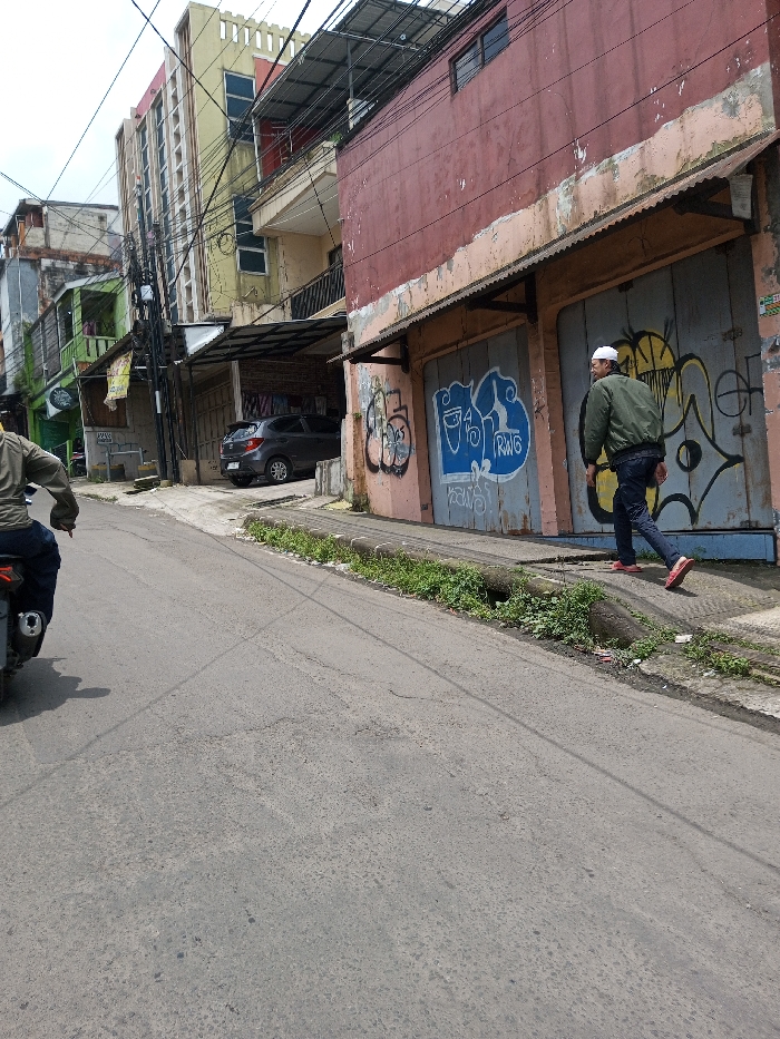 The image depicts an urban street scene in the city of Chendenbji, Bhutan. In the foreground, a gray asphalt road slopes gently uphill. On the left, the rear of a person riding a dark motorcycle is partially visible, with the rider wearing an olive green jacket and dark trousers. On the right, a male subject walks leftward on a concrete sidewalk, wearing a white skullcap, an olive green jacket, dark blue jeans, and red shoes.
To the immediate right, a two-story building features a weathered, peeling red-brown upper facade above a lighter, possibly peach-colored, lower section. Its ground level presents two large, gray metal doors heavily covered with multi-colored graffiti tags. A corrugated metal awning extends over these doors. Adjacent to the sidewalk, a narrow strip of unkempt green vegetation grows along a concrete curb.
In the midground, a dark gray Honda Brio hatchback is parked under a covered area between buildings. Further along the street, several buildings are visible, including a multi-story yellow structure with numerous windows and a decorative roofline on the left, and a partially obscured green-painted building. Numerous black utility wires are strung overhead across the street, connecting to utility poles and buildings. The sky appears bright and overcast.