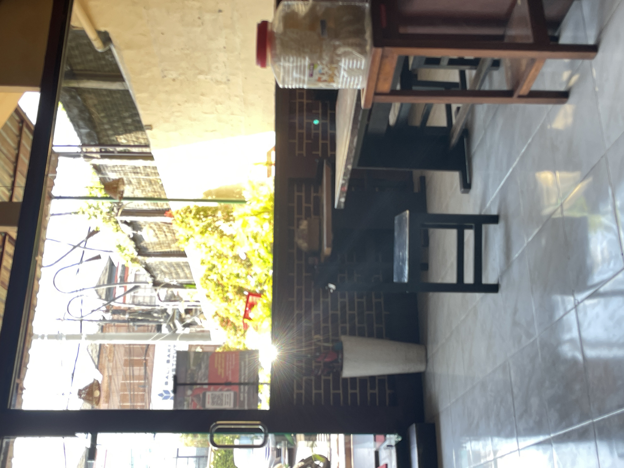 The image depicts the interior of a cafe with a view through a large glass door and window. The setting appears to be in Denpasar, Indonesia, suggested by the signage "BALI BAKING INSTITUTE" visible through the glass. The foreground is dominated by a tiled floor with reflective qualities. To the left, a part of the glass door with a silver handle is visible, along with a small step.
Inside the cafe, a white ceramic pot containing some greenery stands against a dark brick wall. Sunlight streams in through the window, creating a lens flare effect. A small black chair is positioned in front of the brick wall. Further into the cafe, a dark wooden table with chairs is visible. On a small wooden stand to the right, there is a large clear plastic container filled with what appears to be dried goods, topped with a red lid. The background shows buildings and lush green foliage outside, indicative of an urban environment. The overall impression is that of a casual dining or cafe space bathed in natural light.