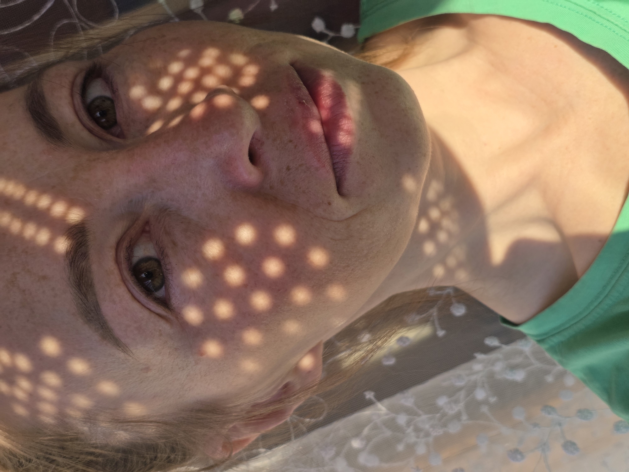 A close-up portrait shows a woman lying on her back with her head tilted slightly. The light source is above and to the left, creating distinct circular patterns of light and shadow on her face, caused by a perforated material. She has fair skin, freckles, and brown eyes. She wears a green shirt. A lace fabric is visible in the lower right corner of the frame. The location is the city of Chaikovsky, Russia.