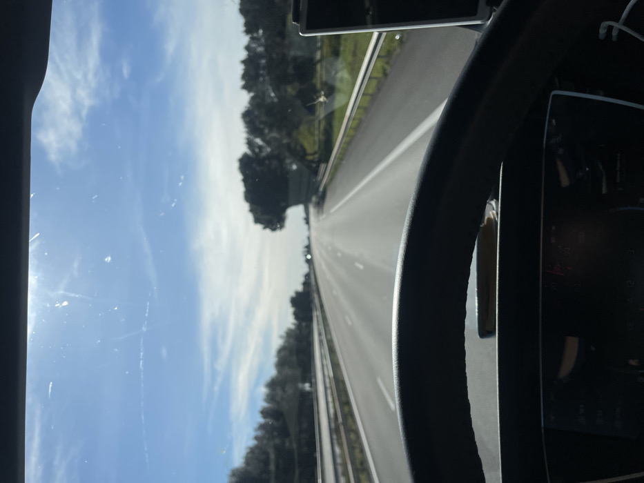 The image depicts a driver's perspective from within a vehicle traveling northbound on a multi-lane highway near Tavel, France, under a clear blue sky with scattered white clouds and a high sun causing lens flare. The asphalt road features white lane markings and stretches straight into the distance, reflecting sunlight. Guardrails line both sides of the highway. To the left, a line of trees and foliage is visible beyond the guardrail. To the right, green vegetation transitions to a cluster of dark green trees, including one distinctively shaped tree. The foreground shows the upper arc of a dark steering wheel. Below the steering wheel, a digital instrument panel displays unreadable icons and segments. An object, possibly a smartphone, rests flat on the dashboard between the steering wheel and the instrument panel. On the far right, a vertically mounted electronic device is partially visible with a white cable extending downwards. The windshield exhibits minor dust and streak marks. No individuals are visible.
