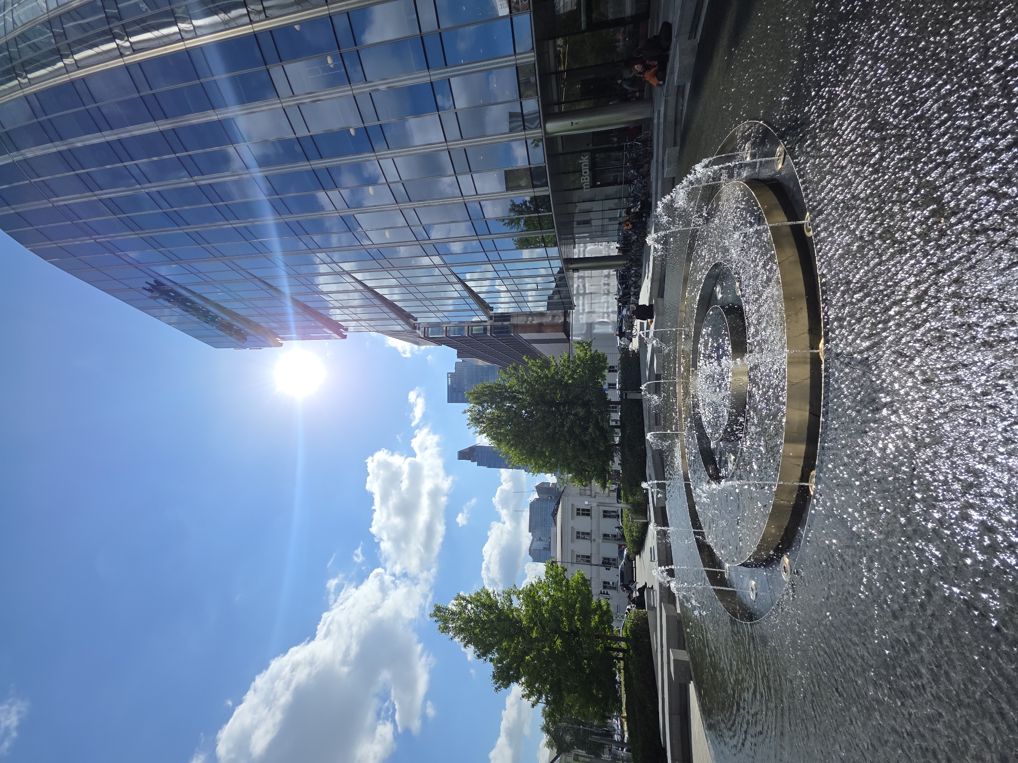 The image captures a sunlit urban scene in Warsaw, Poland, featuring a modern architectural landscape and a water feature. In the foreground, a circular, multi-tiered fountain with golden accents is actively spraying water, creating a shimmering surface. The water reflects the bright sunlight and the surrounding environment. Beyond the fountain, a contemporary glass-fronted building dominates the right side of the frame, its facade reflecting the blue sky and scattered white clouds. A sign on the building reads "mBank". To the left of the building, several mature green trees are positioned, with their foliage catching the light. In the background, other city buildings, including taller skyscrapers, are visible under a clear blue sky with cumulus clouds. The sunlight is intense, creating lens flare in the upper right quadrant of the image. Several individuals are seated on benches or the edge of the fountain area, and bicycles are parked nearby. The scene appears to be a public space, possibly a plaza or courtyard, in a business or commercial district.