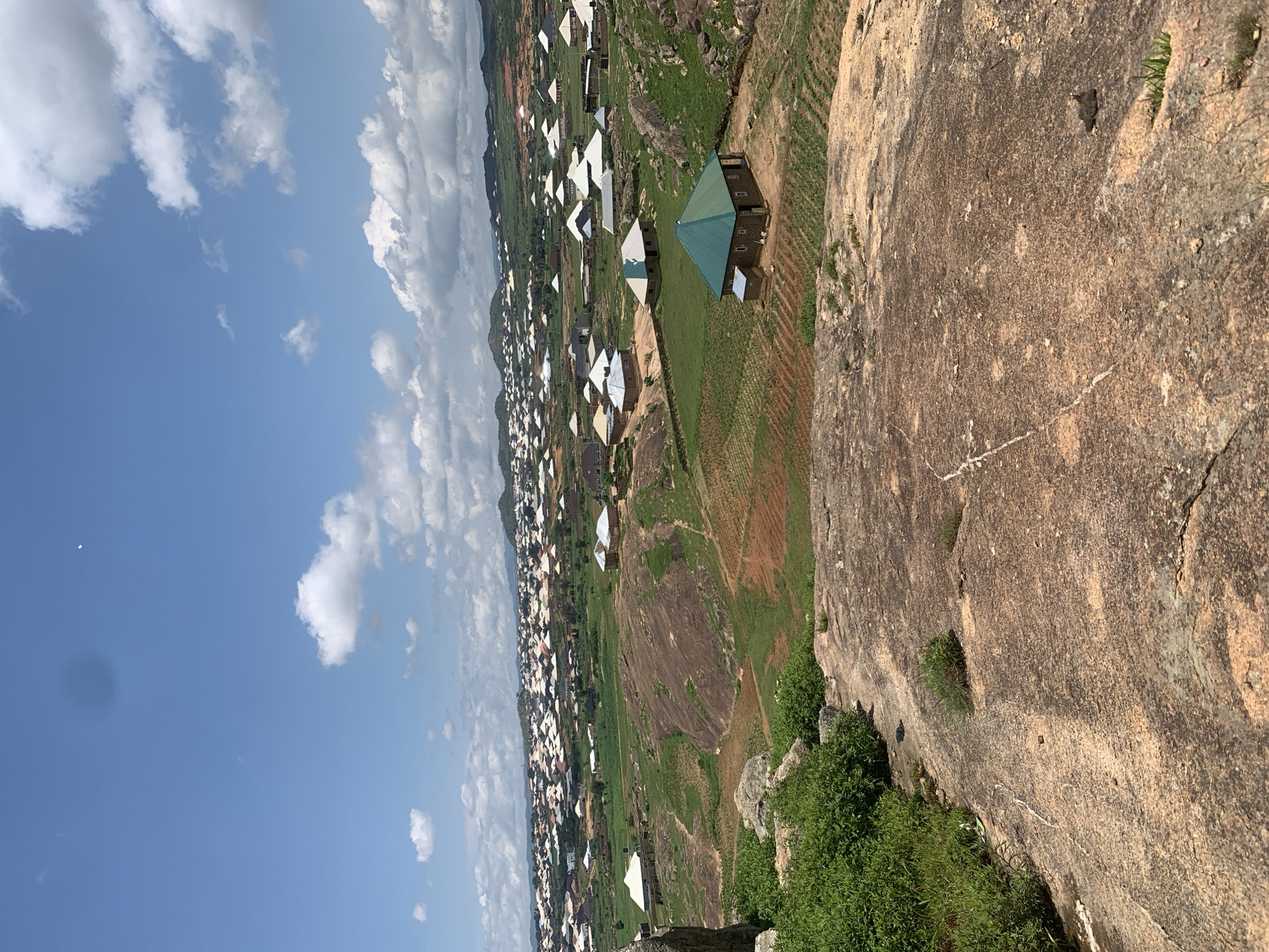 The image presents an elevated, panoramic view of a village in Rafin Bauna, Nigeria, set against a backdrop of rolling hills and a bright blue sky dotted with white clouds. In the foreground, a large expanse of rough, pinkish-brown rock dominates the lower portion of the frame, with patches of green vegetation clinging to its surface.
Beyond the rocky outcrop, the landscape transitions to cultivated fields with rows of young green crops, interspersed with rocky outcrops and patches of dry, reddish soil. Scattered throughout this middle ground are several individual houses, characterized by their simple construction and brightly colored, often white or light-colored, pitched roofs. One prominent house in the middle ground features a vibrant green roof.
Further into the distance, a densely populated residential area stretches across the undulating terrain. This part of the village is a tapestry of houses, mostly with white or similarly light-colored roofs, creating a mosaic against the green and brown of the landscape. The houses appear to be of uniform size and design, suggesting a planned or rapidly developed settlement. In the far distance, more hills, some appearing to be covered in vegetation, form the horizon. The sky above is a clear, vibrant blue with scattered cumulus clouds, some of which are casting faint shadows on the landscape. A sliver of the moon is visible in the upper right quadrant of the sky. No people are visible in the image.
