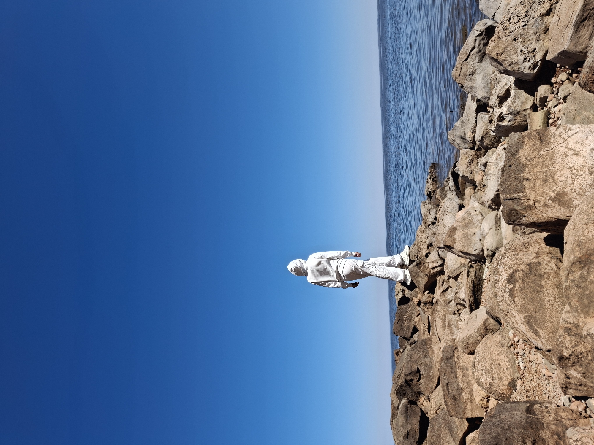 A person wearing a white hooded jumpsuit is standing on large rocks near a body of water, with a clear blue sky in the background. The rocks appear to form a breakwater or shoreline. The location is Noviy, Russia.