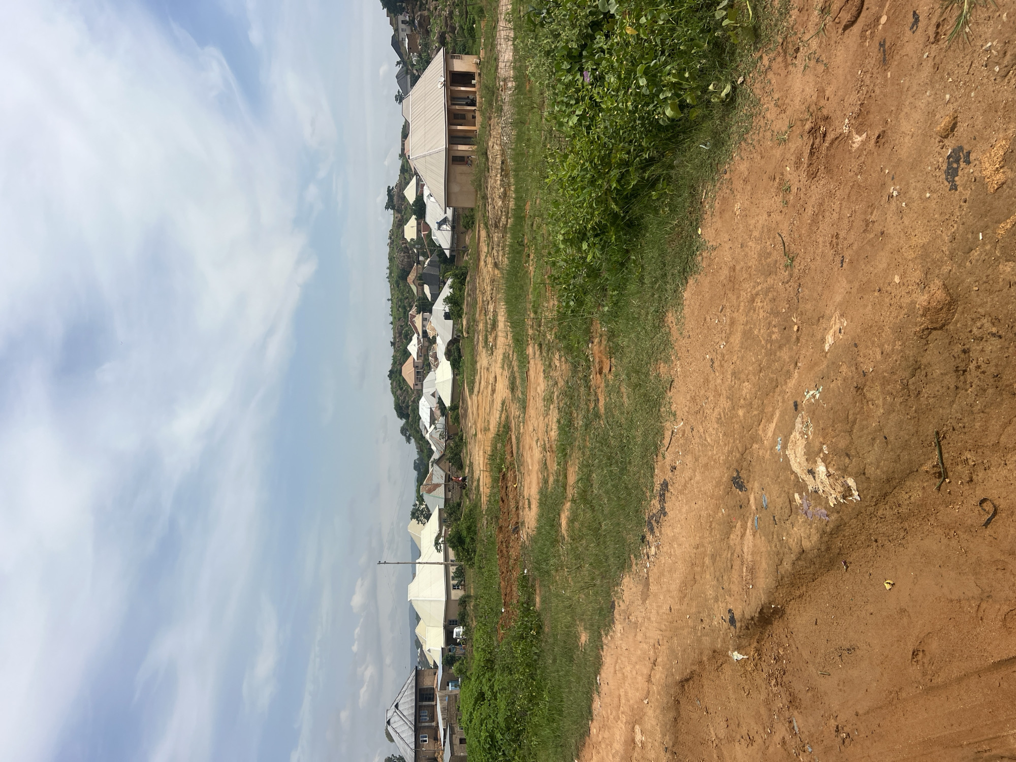 This is a wide-angle, eye-level outdoor shot of a semi-developed residential area in Jos, Nigeria, under a bright, cloudy sky. The foreground is dominated by a reddish-brown dirt road with tire tracks and scattered debris, including small pieces of plastic and what appears to be mud. Green grass and some leafy vegetation grow along the edges of the road and in patches throughout the scene.
In the middle ground and background, a cluster of houses with corrugated metal roofs of various colors, predominantly white and silver, are visible. Some of the houses appear to be newly constructed or under renovation, with unfinished elements and materials present. One prominent house in the mid-right is a single-story building with a light-colored facade and a white, pitched roof supported by pillars. Another larger, multi-story house with a dark, sloped roof is situated on the far left. The terrain rises behind the houses, with rocky outcrops and more vegetation visible on the hills. A single utility pole stands near the center of the image. Two individuals are discernible: one appears to be walking near a white vehicle parked between some houses, and another person is visible further in the distance near a reddish-brown structure. The overall impression is of a community in transition, with ongoing construction and development amidst a natural, hilly landscape.