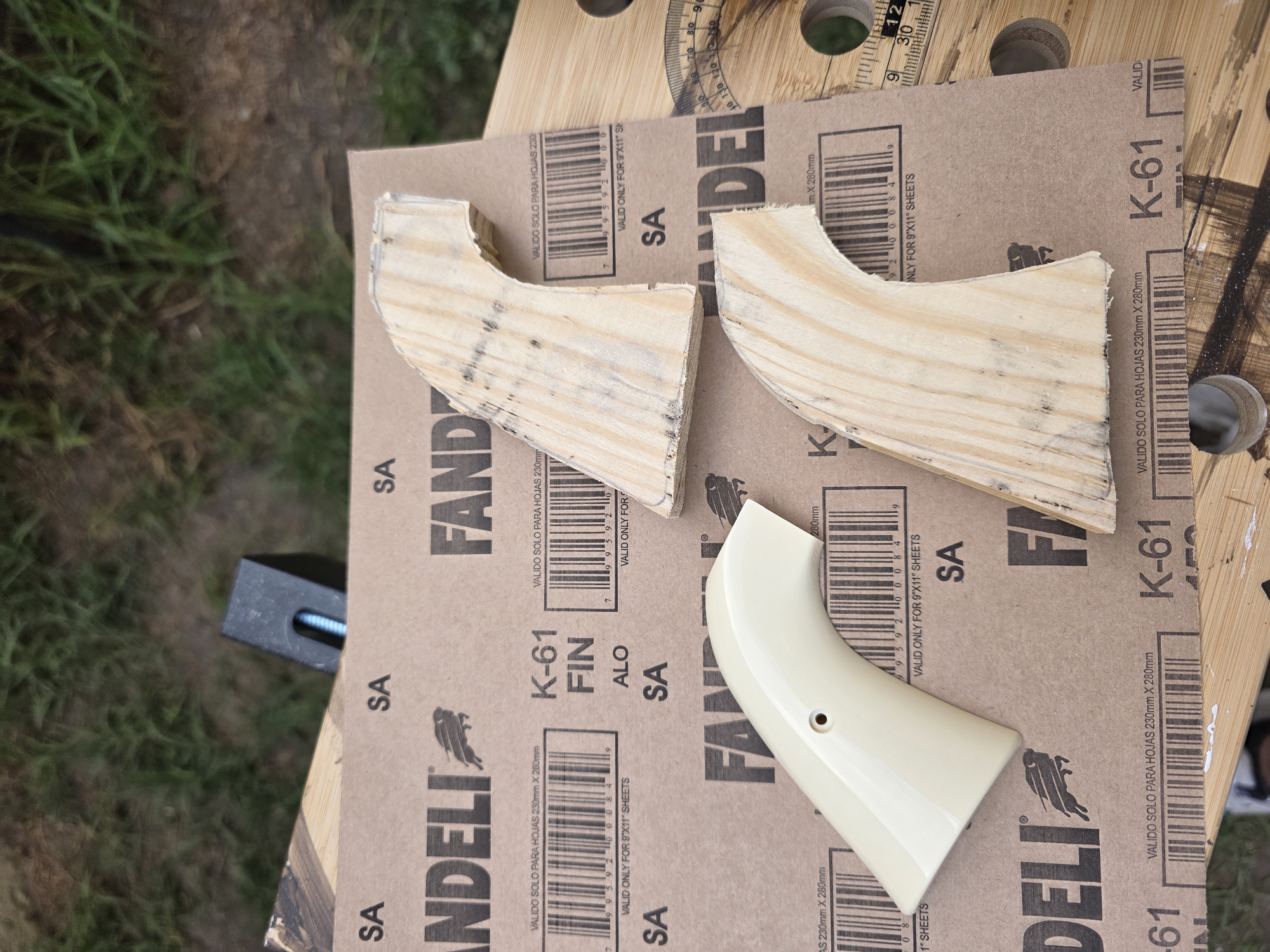 The image shows parts of a handmade gun grip on a workbench. There are two unfinished wooden grip halves showing light wood grain. A third grip half is finished in off-white. The grip parts rest on a sheet of Fandeli brand K-61 sandpaper. The backdrop is a workbench with holes and measuring marks. A clamp is visible at the edge of the workbench. The location is not discernible from the image.