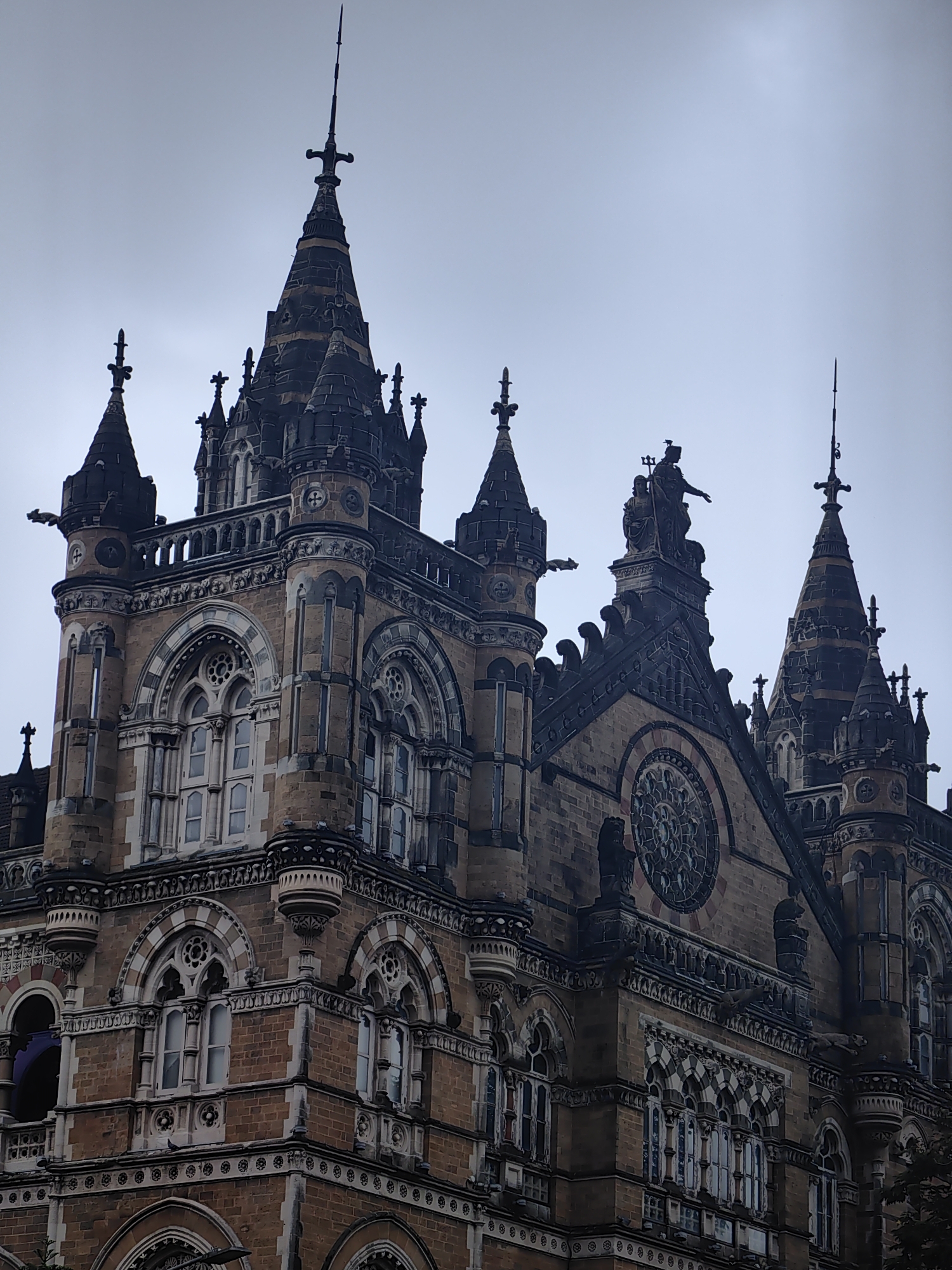 The image depicts the exterior of Victoria Terminus (now Chhatrapati Shivaji Maharaj Terminus) in Mumbai, India. The architectural style is Victorian Gothic Revival, characterized by its intricate stonework, pointed arches, ornate spires, and numerous decorative elements. The building is constructed primarily of sandstone and granite, with the stone exhibiting a warm, earthy hue.
The composition focuses on the upper portions of the structure, highlighting several prominent features:
* **Spires and Pinnacles:** Multiple tall, conical spires and smaller pinnacles cap the building, each topped with finials. The spires are detailed with darker bands and carved embellishments.
* **Gargoyles and Sculptures:** Several grotesque gargoyles protrude from the building's ledges, designed to direct rainwater away. A significant sculpture, depicting a seated figure and a standing figure holding a trident and orb, is visible atop a central peak.
* **Windows:** Elaborate arched windows with intricate tracery are a prominent feature. These windows are multi-paned and appear to be made of glass.
* **Ornamentation:** The facade is richly adorned with decorative moldings, cornices, and carved friezes. Circular windows with stained glass-like patterns are also visible.
* **Sky:** The sky is overcast and a uniform light grey, suggesting a cloudy or hazy day.
The image is a close-up, upward-angled shot that emphasizes the scale and architectural complexity of the building. There are no people or visible signs of contemporary activity in the frame. The overall impression is one of grandeur and historical significance.