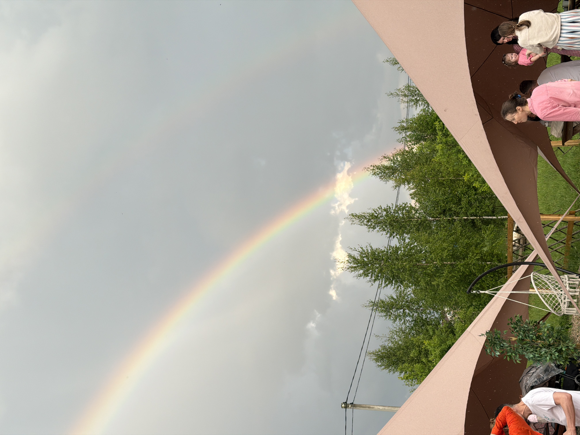 In Zhirnosovo, Russia, a double rainbow arches across a cloudy sky, framing a pastoral scene. Below the celestial display, lush green trees form a backdrop to a gathering of people beneath two large, pale pink canopies. On the left, a man in a white t-shirt is partially visible, as is a stroller and a decorative plant. To the right, under the other canopy, a group of individuals are conversing. A woman in a pink shirt is in the foreground, facing away from the camera. Behind her, a woman holds a young child dressed in pink. Further back, another woman in a cream jacket and striped skirt stands with her back to the viewer, engaged in conversation. A white macrame swing chair hangs nearby, adding to the relaxed atmosphere.