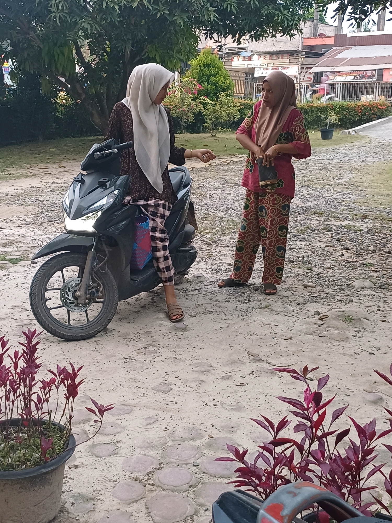 Two women are standing outdoors in a sandy area with scattered paving stones. The woman on the left is seated on a dark-colored motorcycle, wearing a patterned brown shirt and checkered pants. She has a light-colored hijab and is wearing brown sandals. A patterned bag is hanging from the side of the motorcycle. The woman on the right is standing and wearing a pink patterned shirt and matching pants. She has a brown hijab and is holding a dark-colored bag. The background shows trees, buildings, and a fence, suggesting an urban or semi-urban setting in Dumai City, Indonesia. The overall lighting suggests it is daytime.