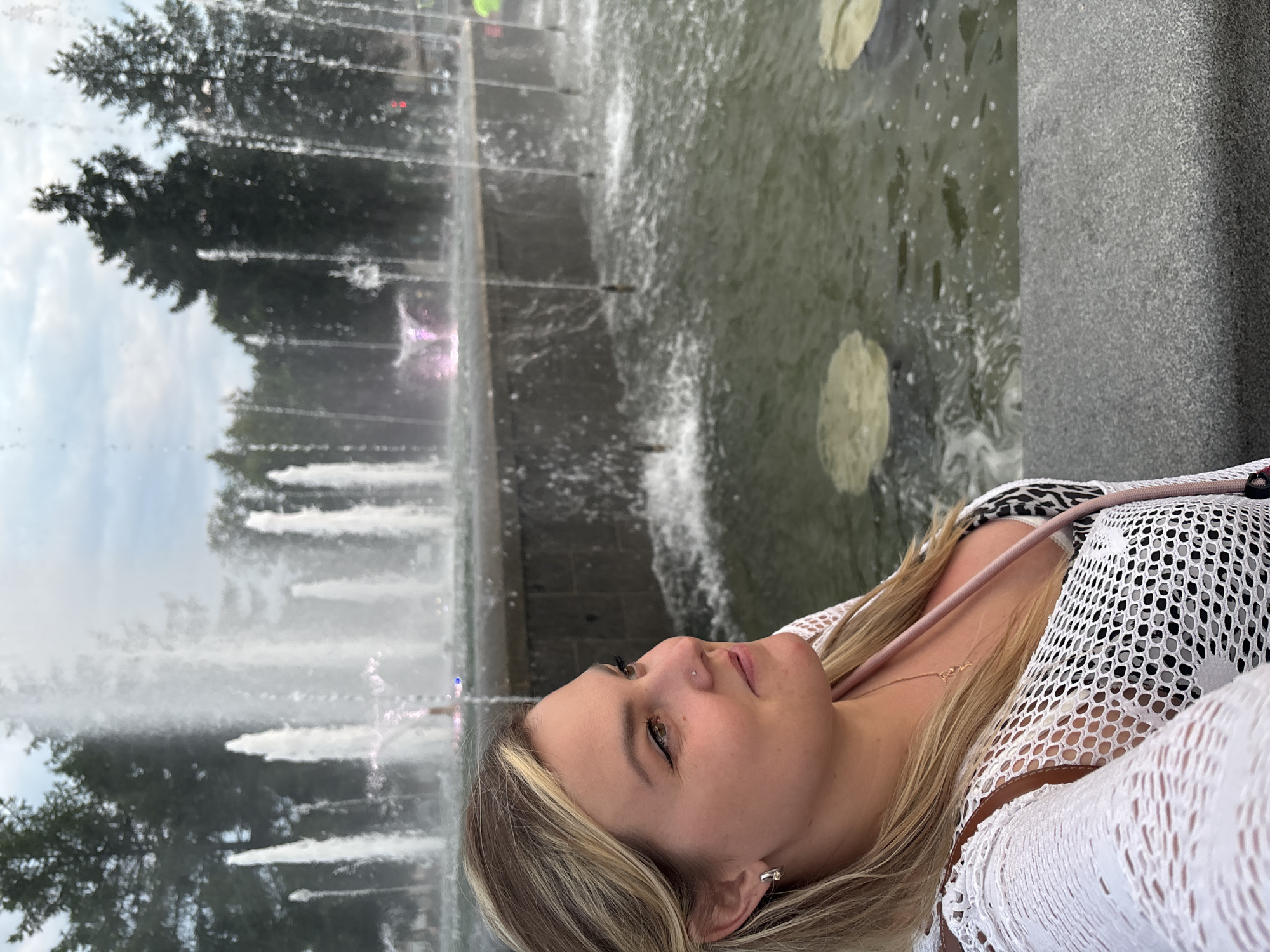 A woman with blonde hair, wearing a white mesh cover-up over a black and white patterned top, is looking to her right in front of a large fountain in Chaikovsky, Russia. The fountain has multiple water jets spraying upwards, some with pink lighting at their base. The water creates a mist that partially obscures the background trees and sky. The woman is wearing earrings and a necklace. She appears to be taking a selfie or is positioned for a photo. The foreground shows a stone ledge and the water of the fountain.