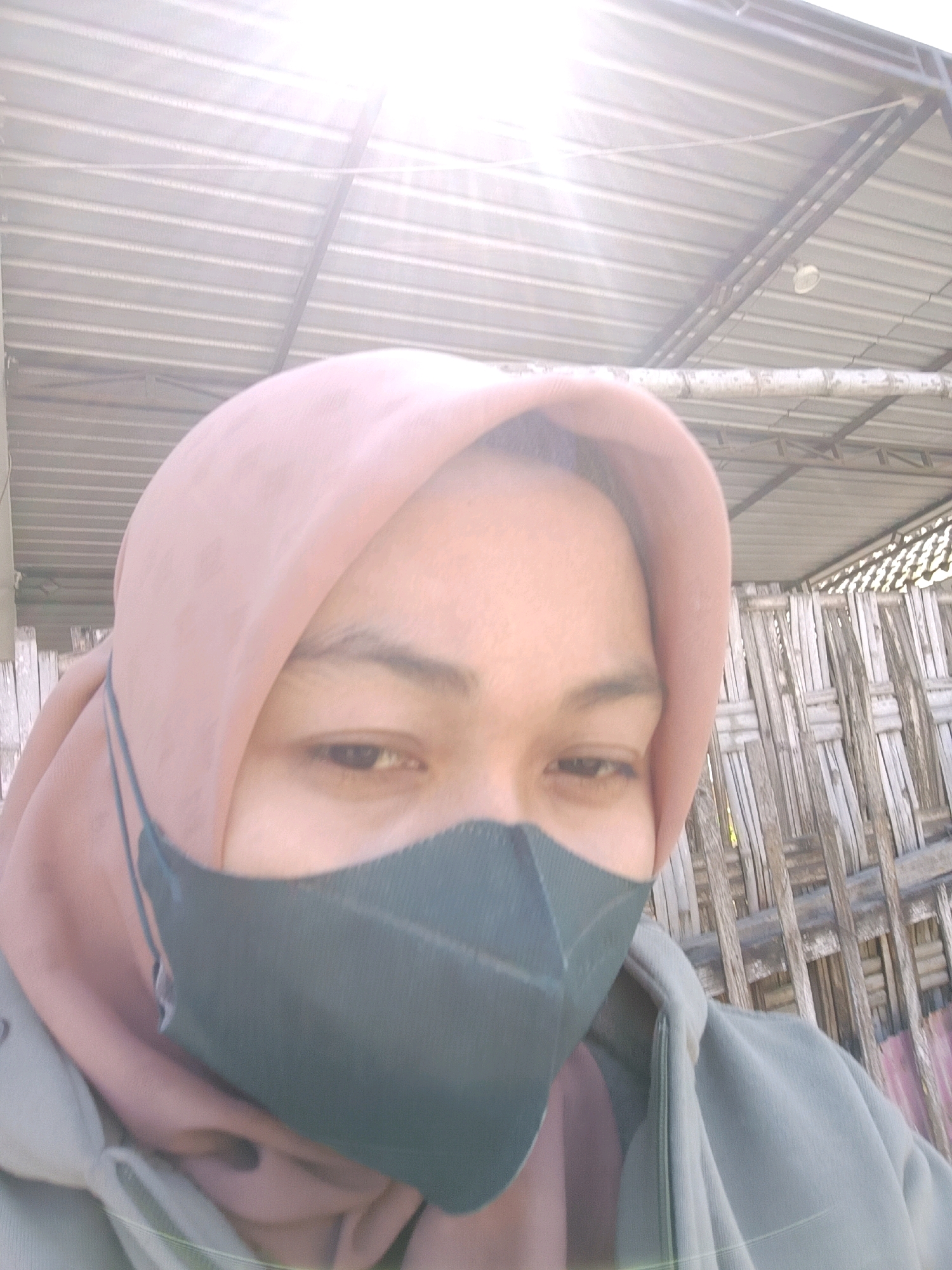A close-up selfie shows a person wearing a pink hijab and a dark gray face mask. They are wearing a light gray hooded jacket. The background features a corrugated metal roof with a thin wire running across it, and a woven bamboo fence. The overall lighting suggests an outdoor setting, possibly during the daytime. The location is identified as Blega, Indonesia.