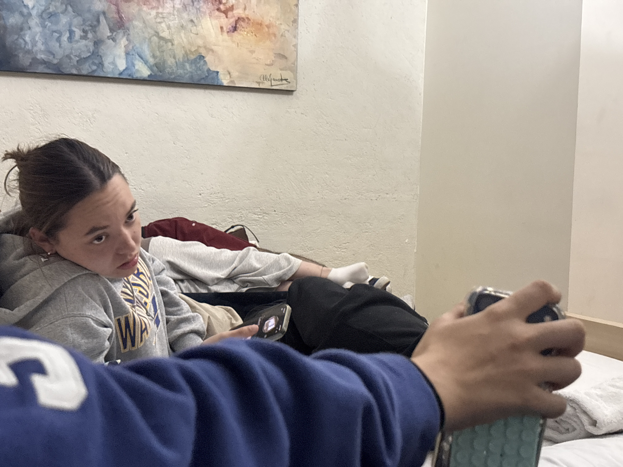 A woman with brown hair pulled back in a ponytail is reclining against a wall. She is wearing a gray hooded sweatshirt with blue and gold lettering. She is holding a smartphone and looking slightly to the left. Several articles of clothing are piled around her, including a gray sweatshirt, a red shirt, and black pants. A pair of feet wearing white socks is visible in the background. In the foreground, a person wearing a blue sweatshirt is holding a smartphone. A painting hangs on the wall behind the woman. The scene is likely inside a room. The location in the city of Puebla, Mexico, cannot be determined from the image.
