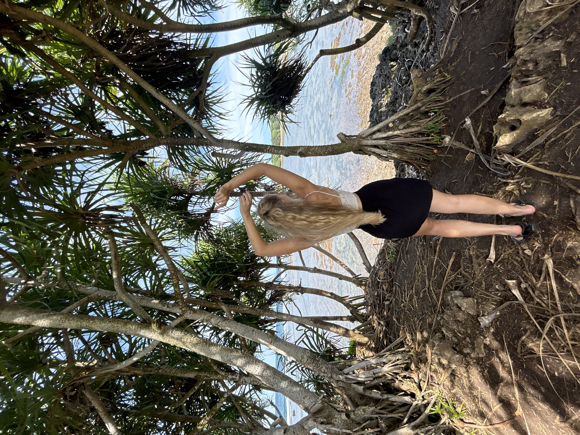 A woman stands among exposed roots of a tree at a beach, looking out over the water. She is wearing a white tank top and a black skirt, and sandals. Her blond hair cascades down her back. The image is taken from a low angle, looking upward, and the tree branches and foliage fill much of the frame, partially obscuring the view of the ocean. The location is near the coastline in Kuta Selatan, Bali, Indonesia. Exposed roots and dark soil are near her feet.
