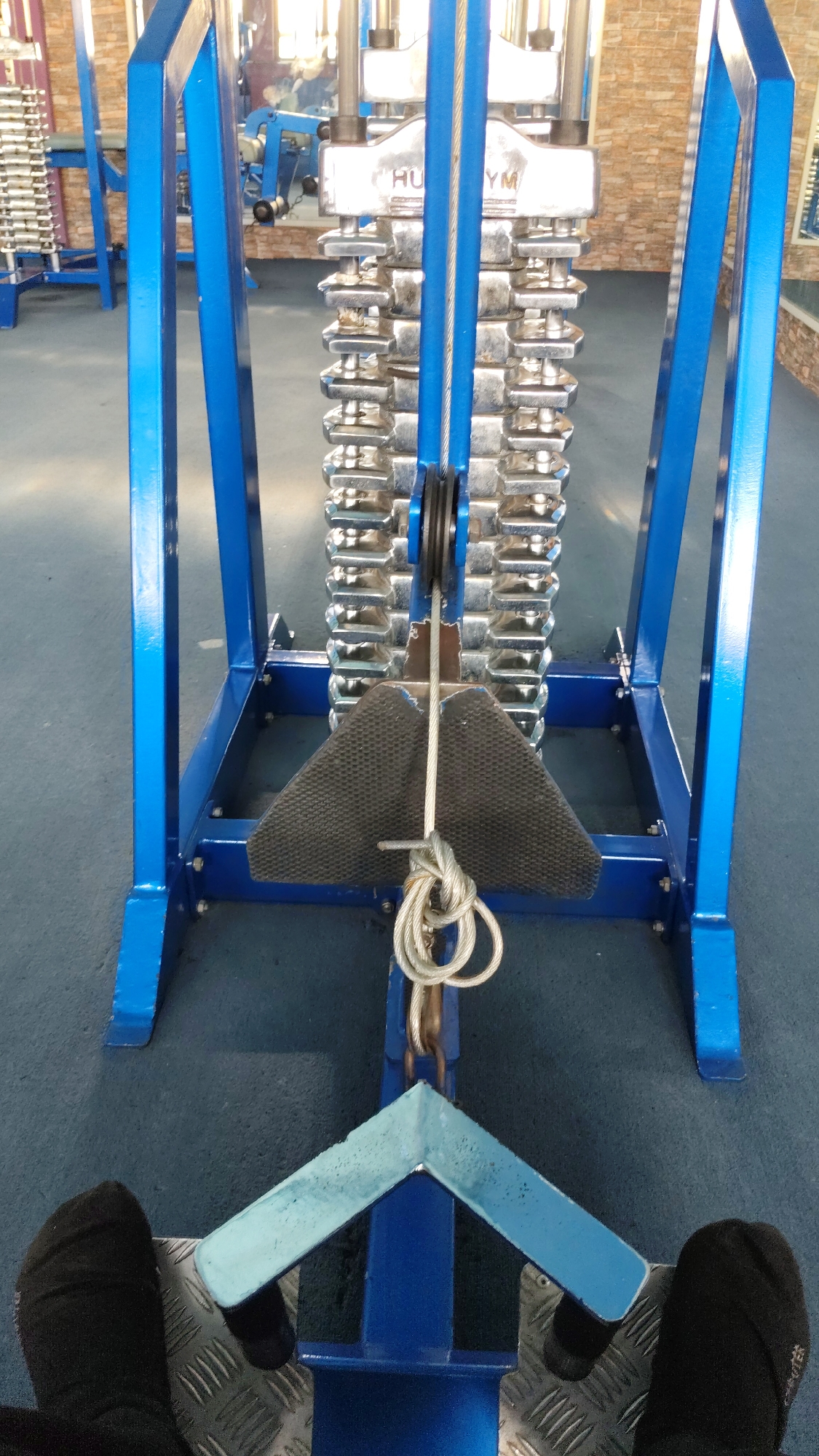 The image shows a close-up view of a weight stack machine in a gym. The machine has a blue metal frame and a stack of silver weights. A cable runs from the top of the weight stack down to a black triangular footplate. The scene is viewed from the perspective of someone sitting on the machine, with their black-socked feet visible on metal footrests in the foreground. The gym floor is a dark blue carpet. The background is blurred, but appears to show other gym equipment and a brick wall. There is no specific indication of the location being Munggut, Indonesia, other than the provided information that it is the city of Munggut, Indonesia.