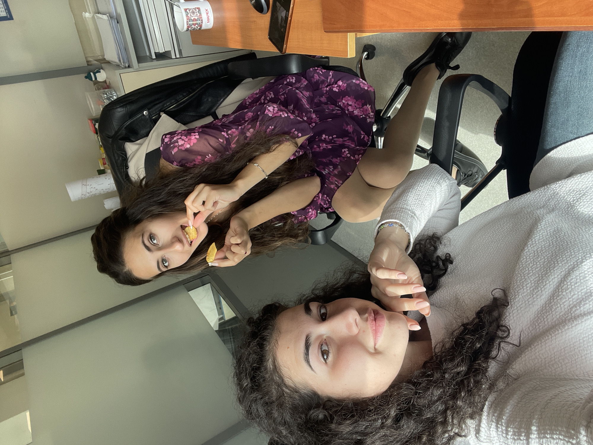 This high-angle, selfie-style photograph captures two young women in what appears to be an office or study environment in Sofia, Bulgaria. The image is rotated counter-clockwise, making the subjects appear sideways.
The woman in the foreground has long, dark, curly hair and is wearing a white textured top or sweater. She looks directly at the camera with a neutral expression, her right hand with light pink manicured nails held near her face. A colorful beaded bracelet is visible on her left wrist.
Behind her, another woman with long, dark, straight hair is seated in an office chair, wearing a purple dress with a pink floral pattern. She holds two pieces of food, resembling chips or crisps, one near her mouth and another in her hand, suggesting she is eating. She also looks towards the camera with a slight smile. She wears black shoes and a silver bracelet on her left wrist.
The setting includes a wooden desk at the top right, holding stacked papers, books, and a mobile device or laptop. A white mug with the word "MAPEX" and a decorative pattern is clearly visible on the desk. A black leather jacket is draped over the back of a chair. The background features a light-colored wall and a glass partition, characteristic of an indoor office or communal space. Bright natural light indicates it is daytime. Both women appear relaxed and engaged in a casual moment.