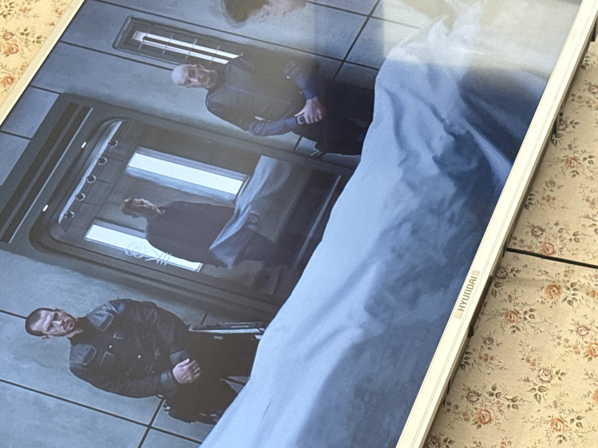 The image appears to be a photo of a screen displaying a scene. The scene shows two men in what appears to be a corridor or room of some kind, standing beside what is likely a covered body on a gurney.
Location: Unverifiable; no discernible information suggests Mytishchi, Russia.
People:
- Two men, both fair-skinned. One is bald; both wear dark clothing.
Subjects:
- An object covered by a blue sheet, likely a body on a gurney.
Objects:
- A gurney
- A blue sheet
- A screen (likely a television)
- A light fixture
- A door or entrance
Actions:
- The men are standing near the gurney.
Interactions:
- The scene suggests that the men might be involved in a formal process related to a body.
Environment:
- A corridor or room.
- Walls are grey.